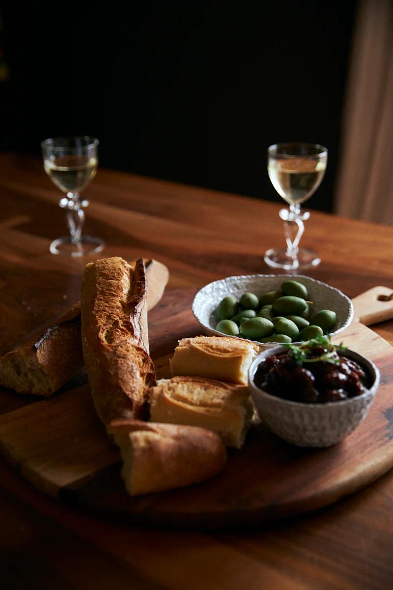 Wooden board with baguette, bread slices, olives, and herb-garnished dip. Mostarda relish and wine glasses on table. Rustic setting.