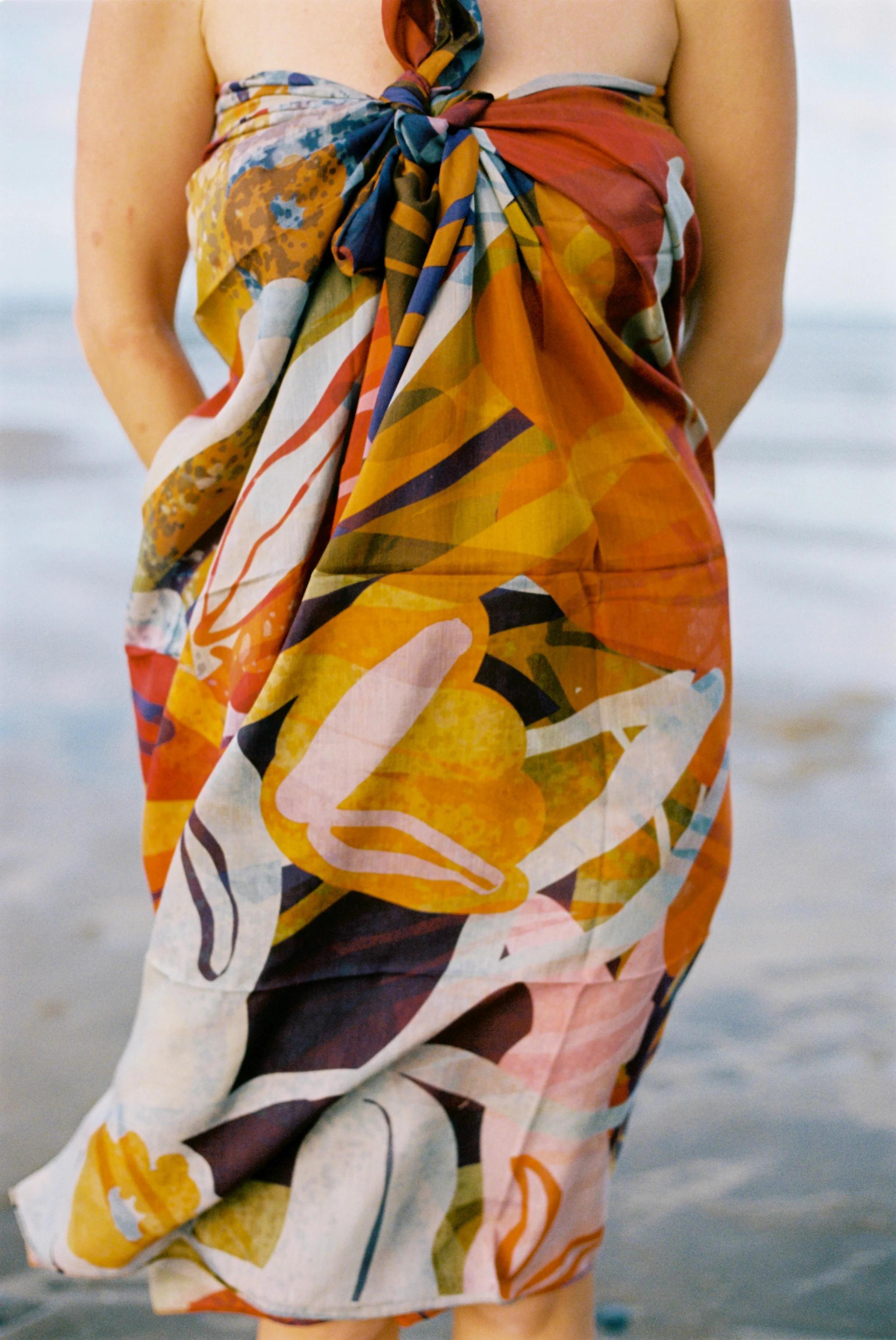 Person in colourful abstract-patterned sarong facing the camera. On beach with water and sand in background.