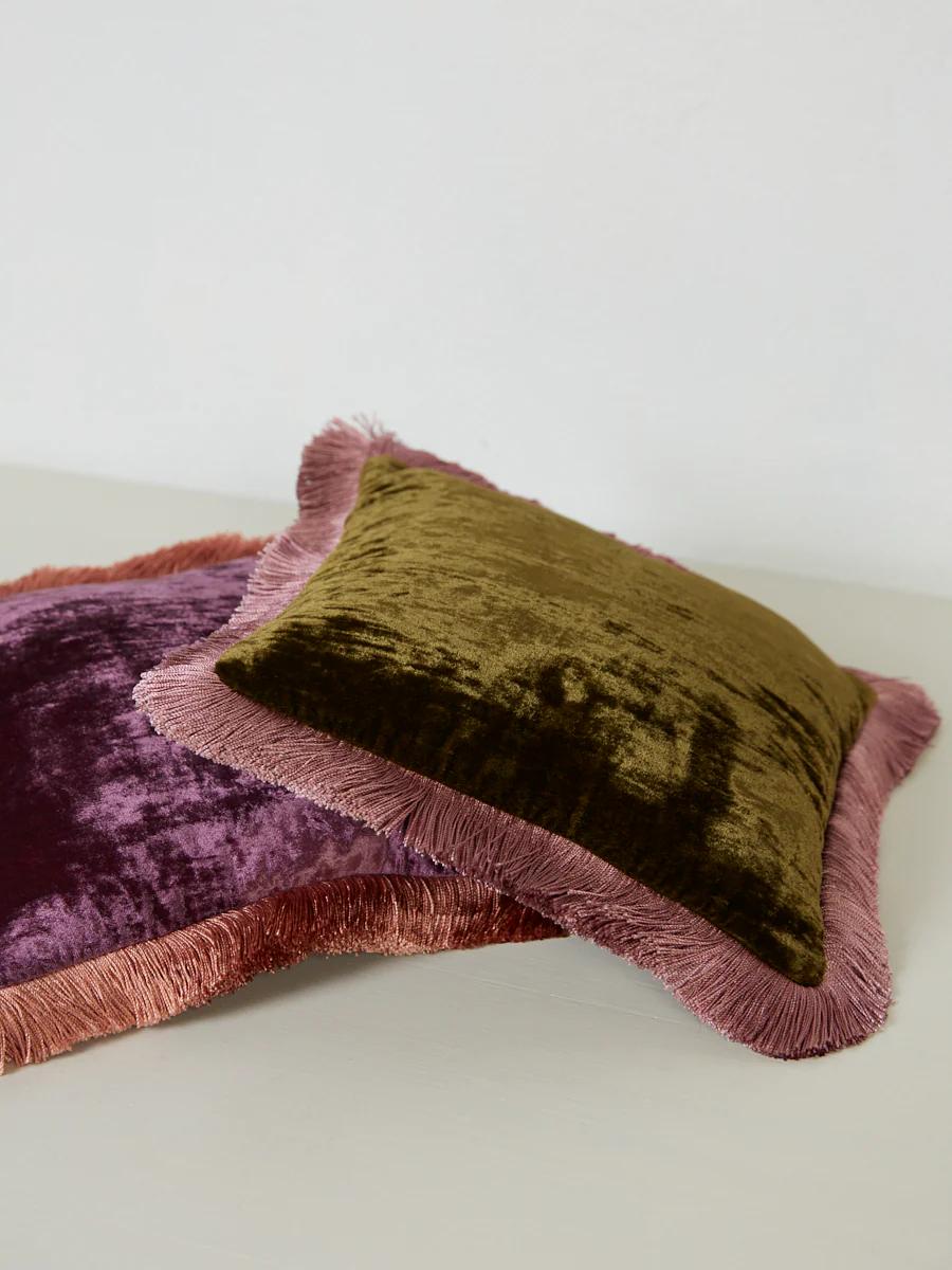 Two velvet cushions with fringed edges are stacked together. The top cushion is olive green, while the bottom is dark purple, both featuring contrasting pink fringes.