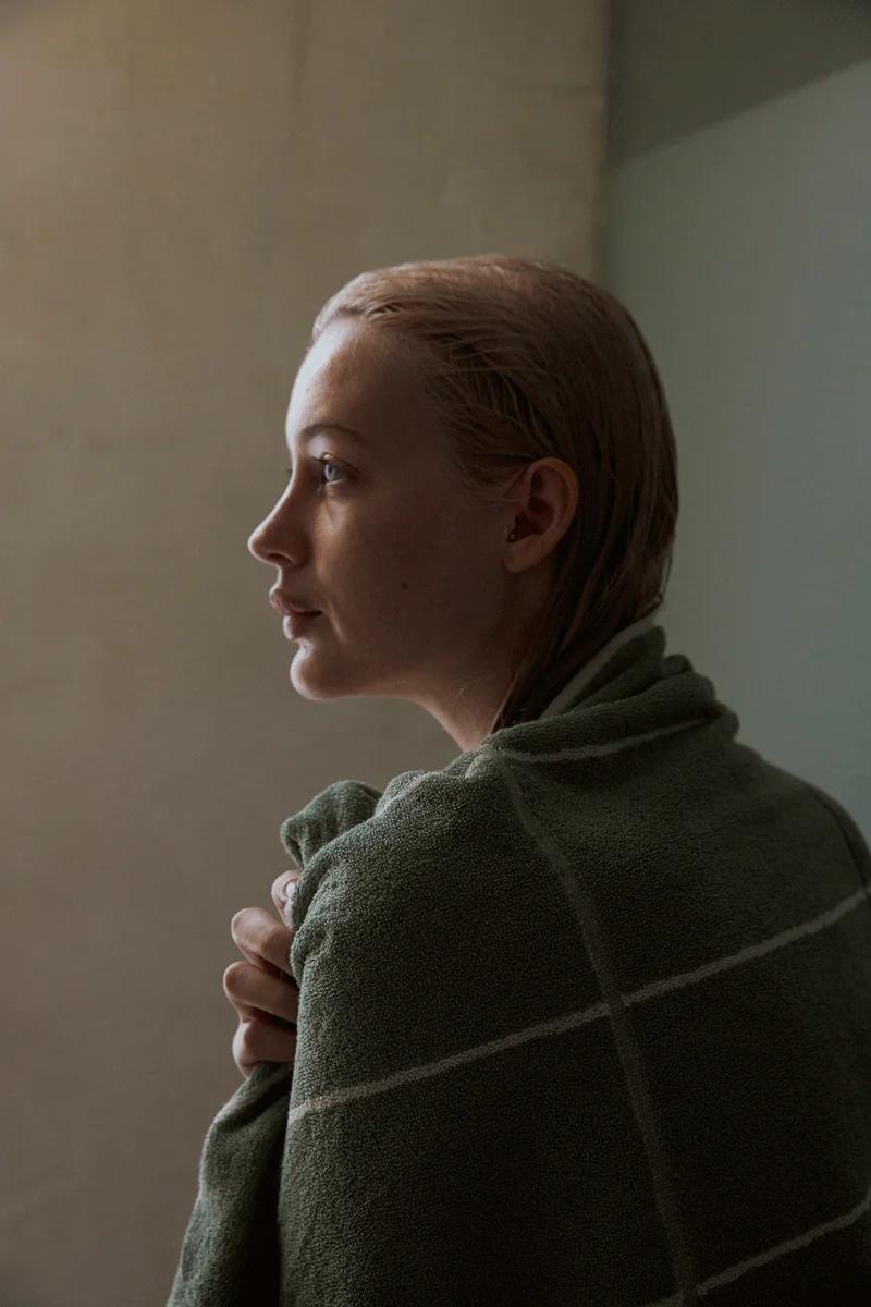 A person wrapped in a green checkered organic cotton towel looks to the left. Soft lighting highlights the towel’s quality against a plain wall, emphasizing their bathing ritual.