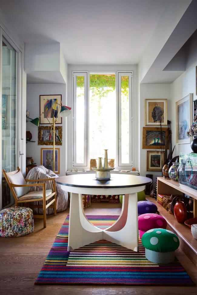 Bright room with round table, mushroom stools, striped rug, and art pieces. Wooden chair with floral cushions near large windows.