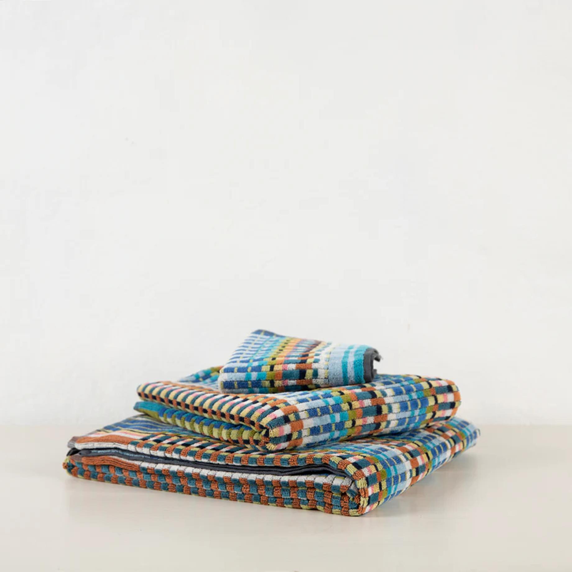 Stacked multicoloured Missoni-style towels with geometric pattern. Blues, greens, oranges, and whites on white surface against light wall.