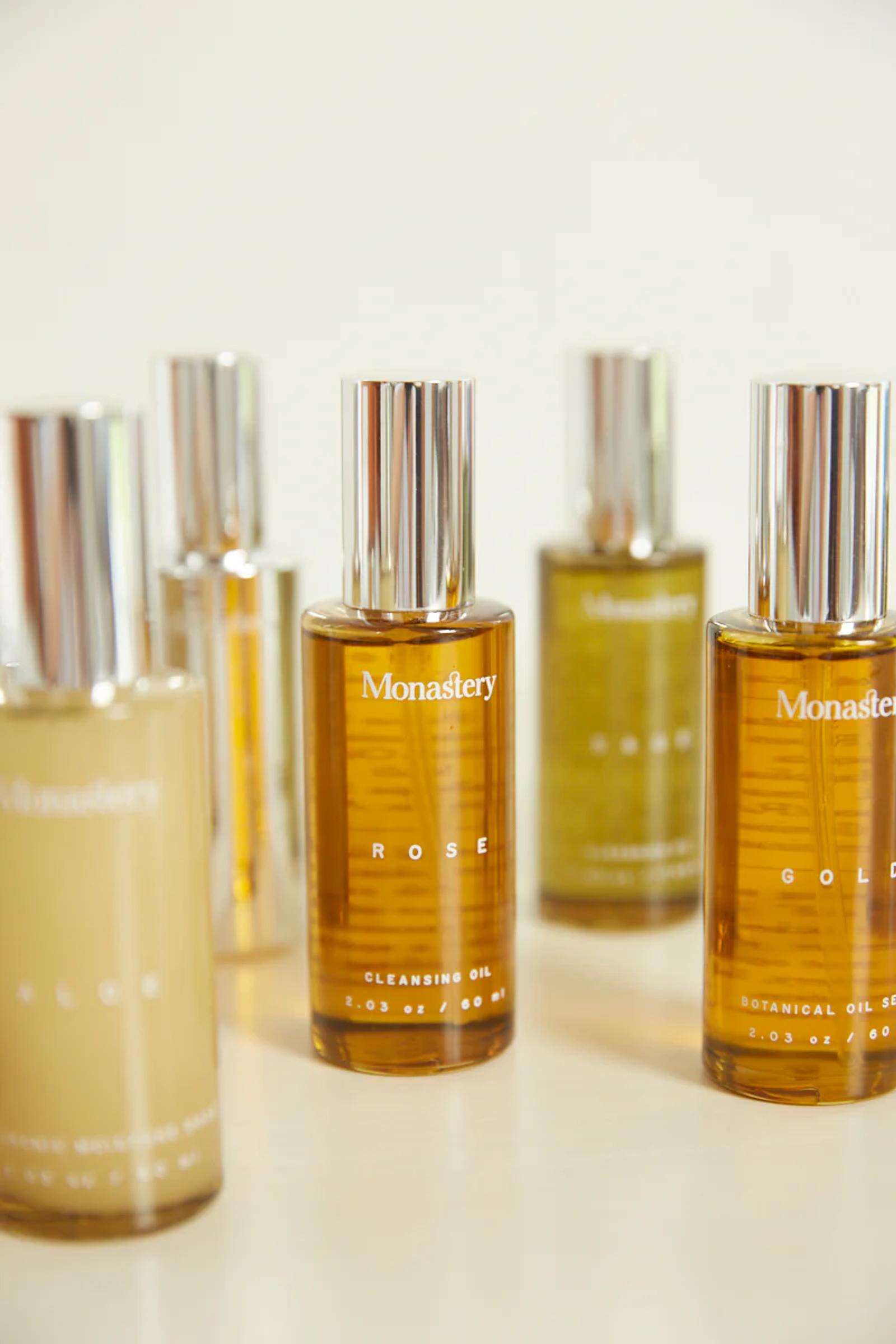 Monastery skincare products displayed, including rose cleansing and gold botanical oils with silver caps. Arranged on light surface with soft background.