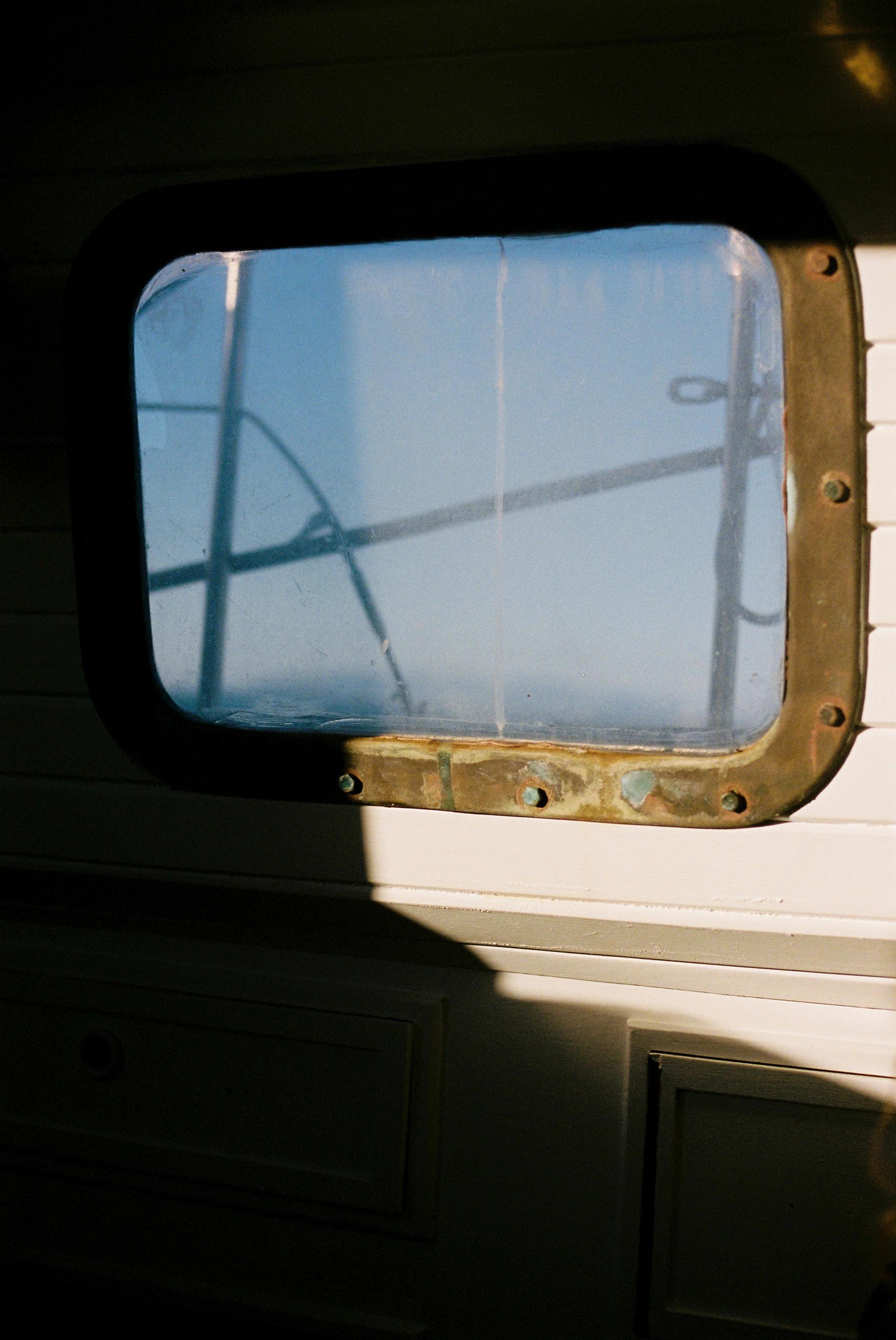 Boat window with worn metal frame. Rigging against blue sky. Sunlight casts shadows inside.