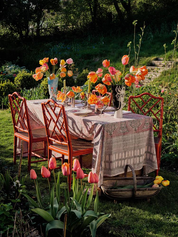 Outdoor table with floral cloth and red chairs. Tulip arrangements on table, surrounded by lush garden.