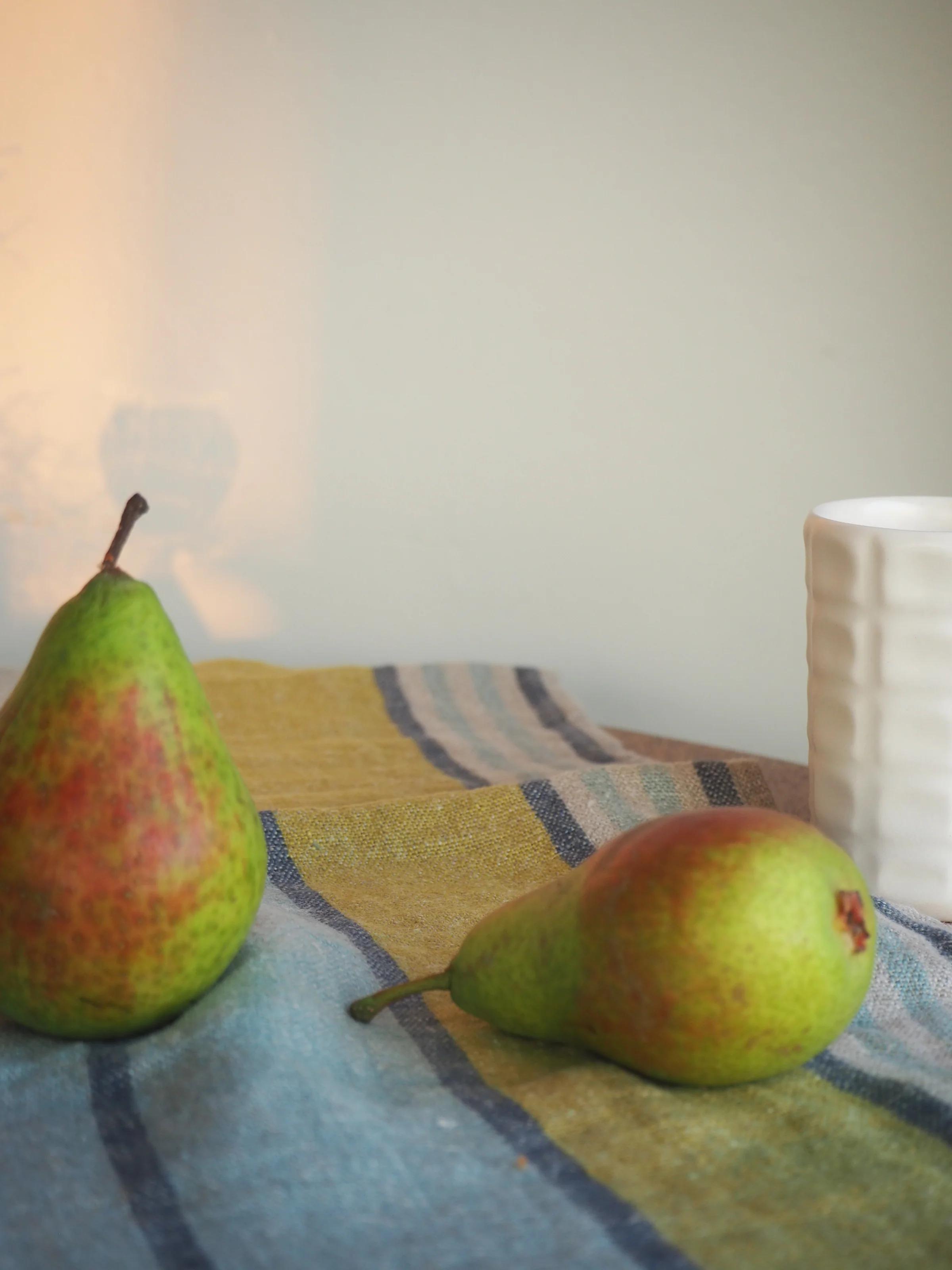 Two green pears on a striped cloth with a white ceramic cup slightly out of focus, evoking a calm and reflective morning mood.