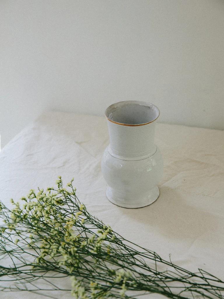 Light grey Astier de Villatte vase on cream tablecloth. Sprigs of small white flowers with green stems lie nearby, adding natural touch to minimalist scene.