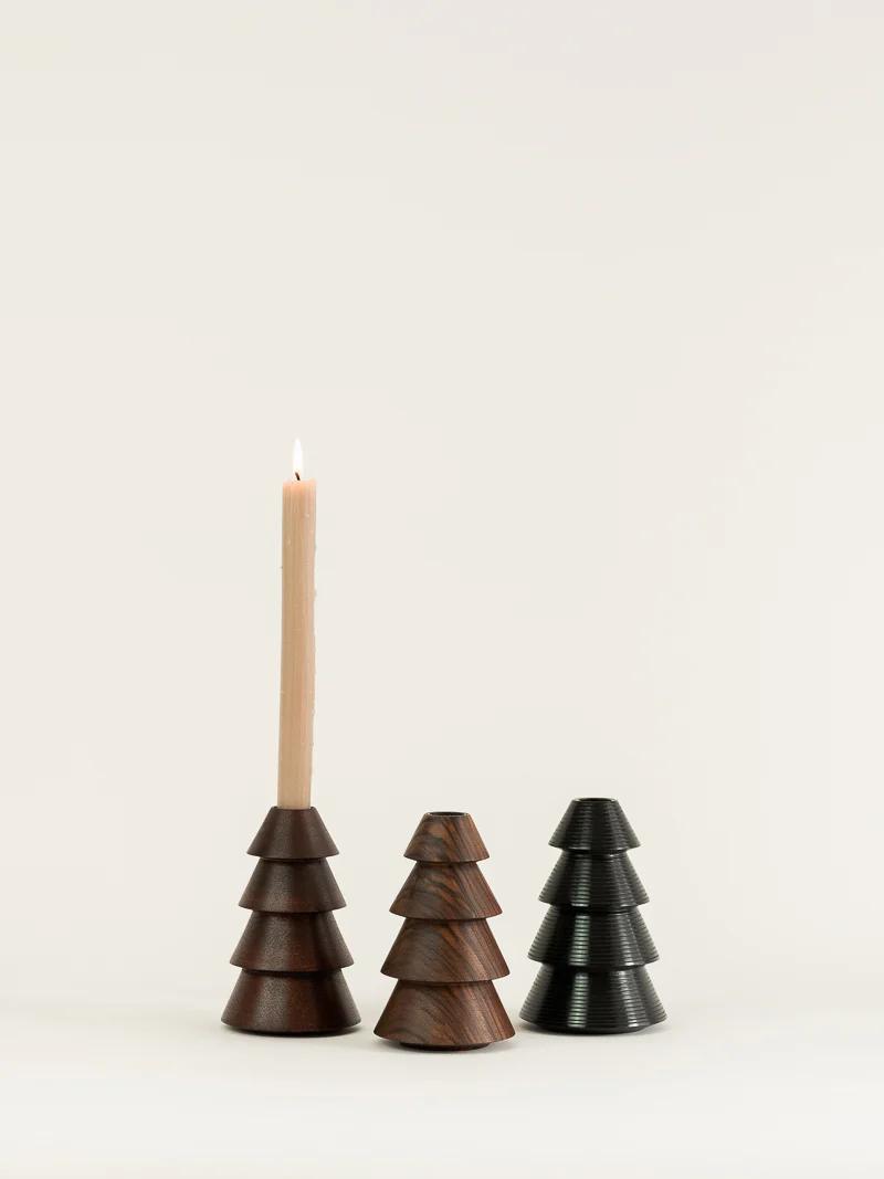 Three pyramid-shaped wooden candle holders are side by side. The leftmost holds a lit candle, while the others are empty. Crafted in a woodturning studio, they vary in colour from dark brown to black with tiered, tree-like designs.