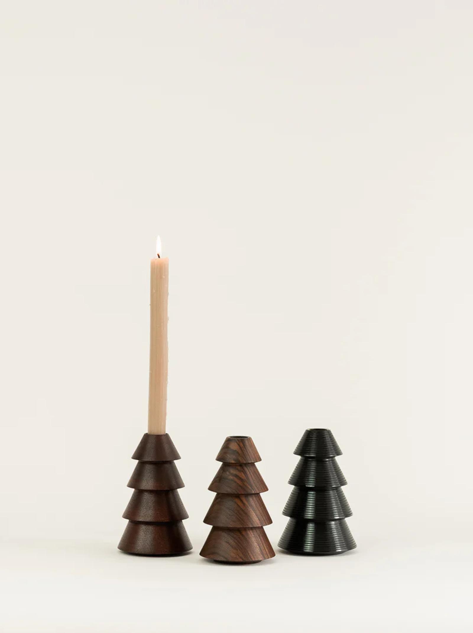 Three pyramid-shaped wooden candle holders are side by side. The leftmost holds a lit candle, while the others are empty. Crafted in a woodturning studio, they vary in colour from dark brown to black with tiered, tree-like designs.