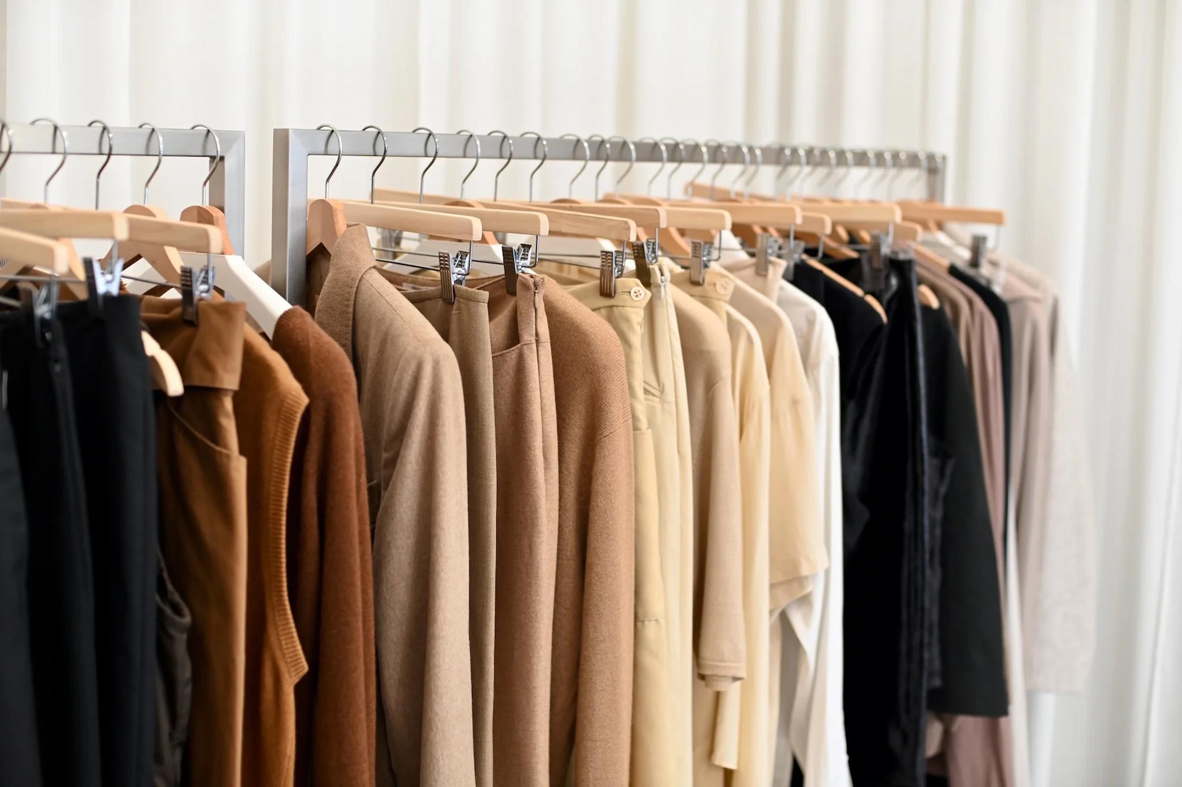 Clothing rack with shirts, jackets, and coats in brown, beige, and black. White curtains in background.