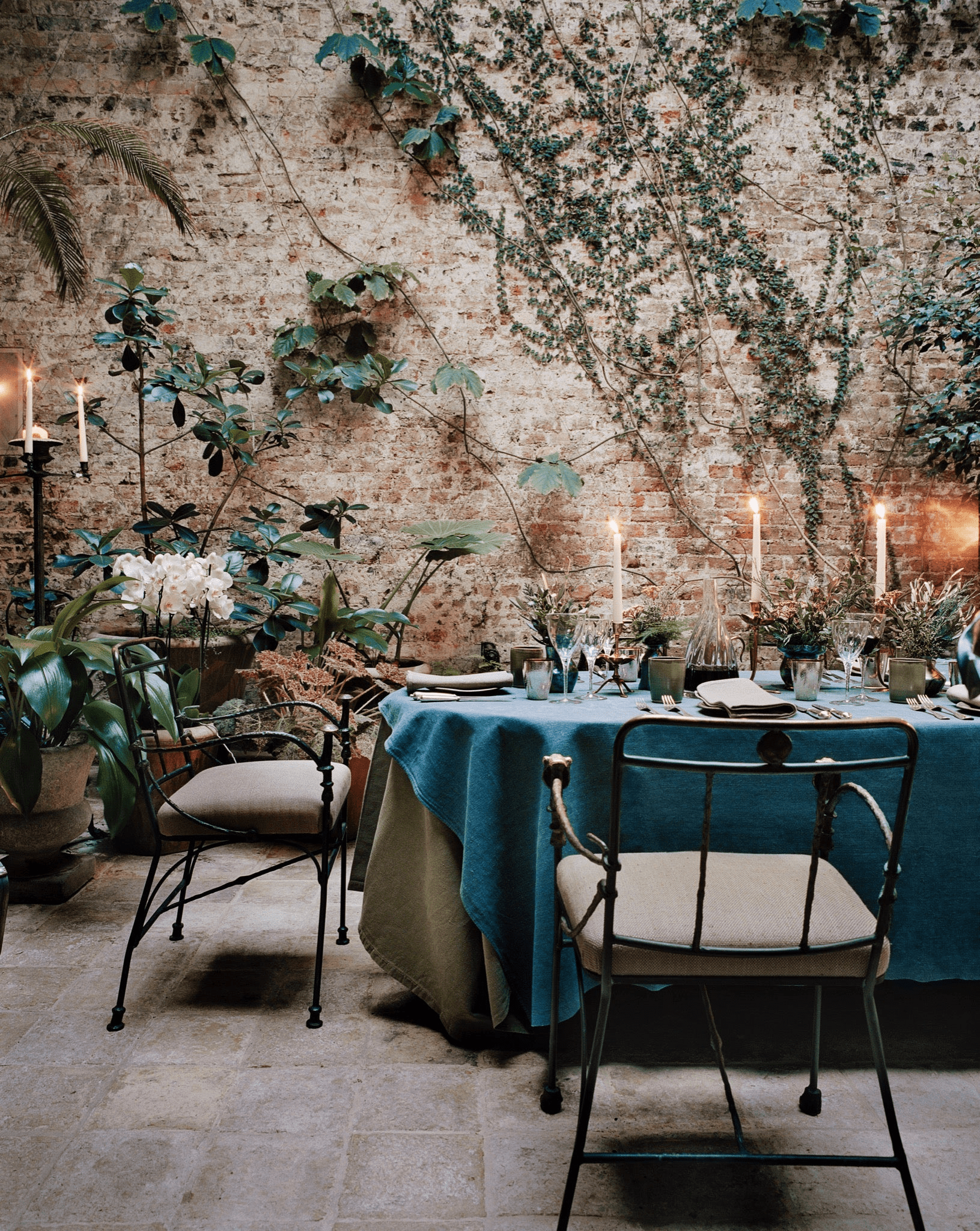 Outdoor dining area with clothed table and metal chairs. Candles, plants, and ivy on brick wall.