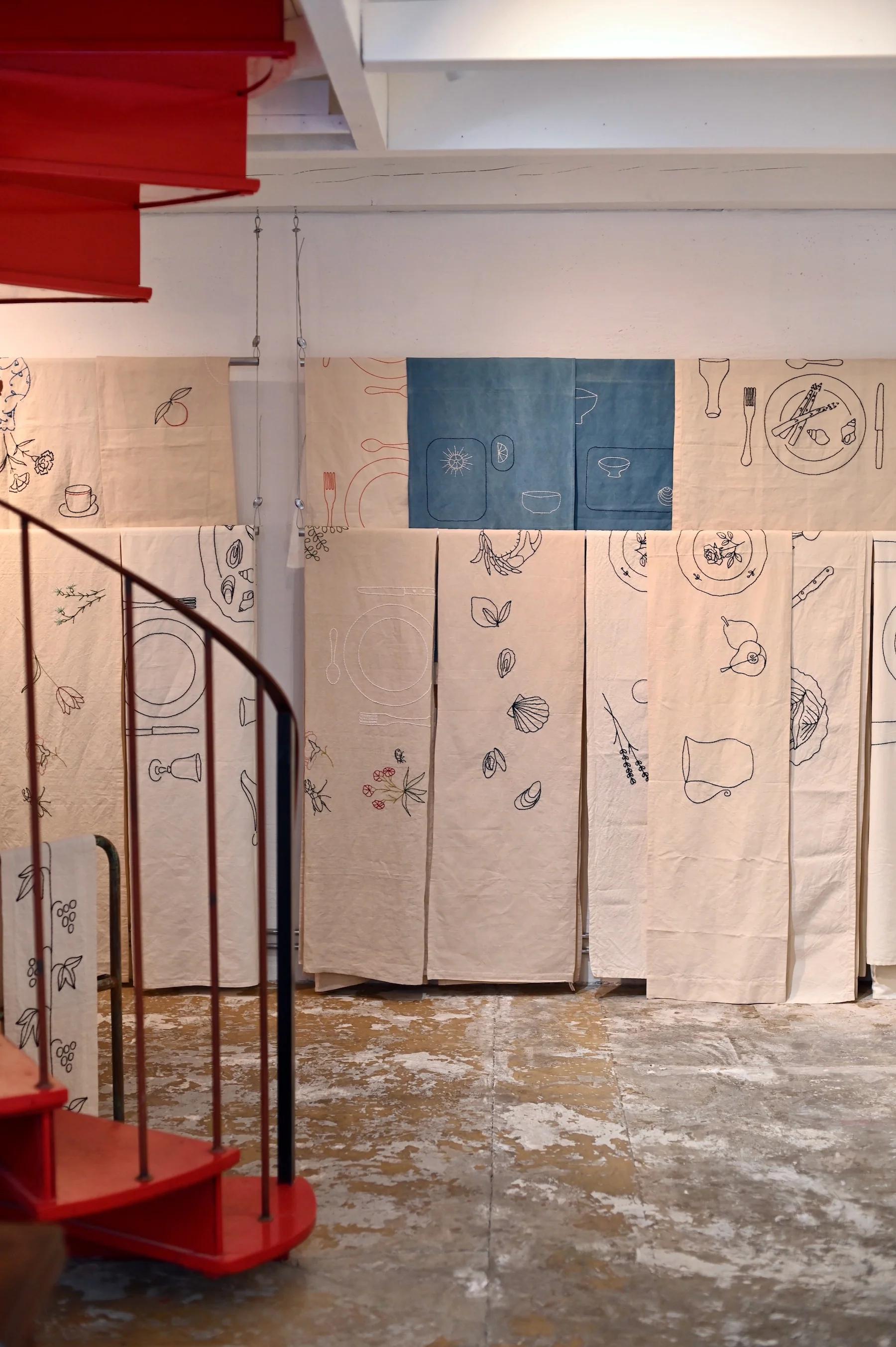 Room with red spiral staircase. Fabric panels with line-drawn kitchen items hang in background. Worn cement floor.