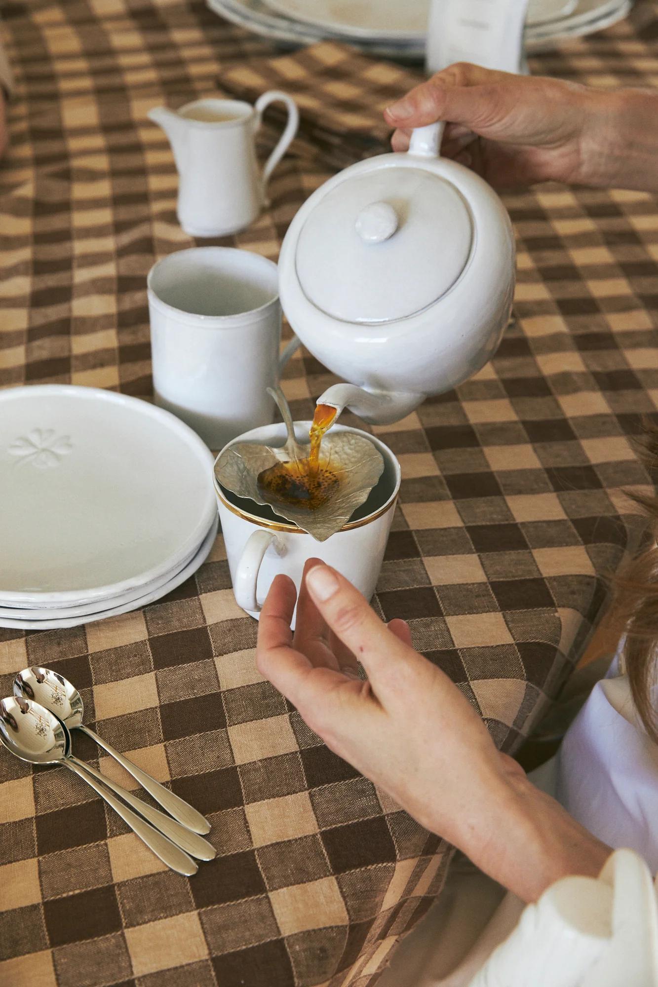 Person pouring tea through filter on checkered tablecloth. Table set with plates, mug, creamer, silverware.