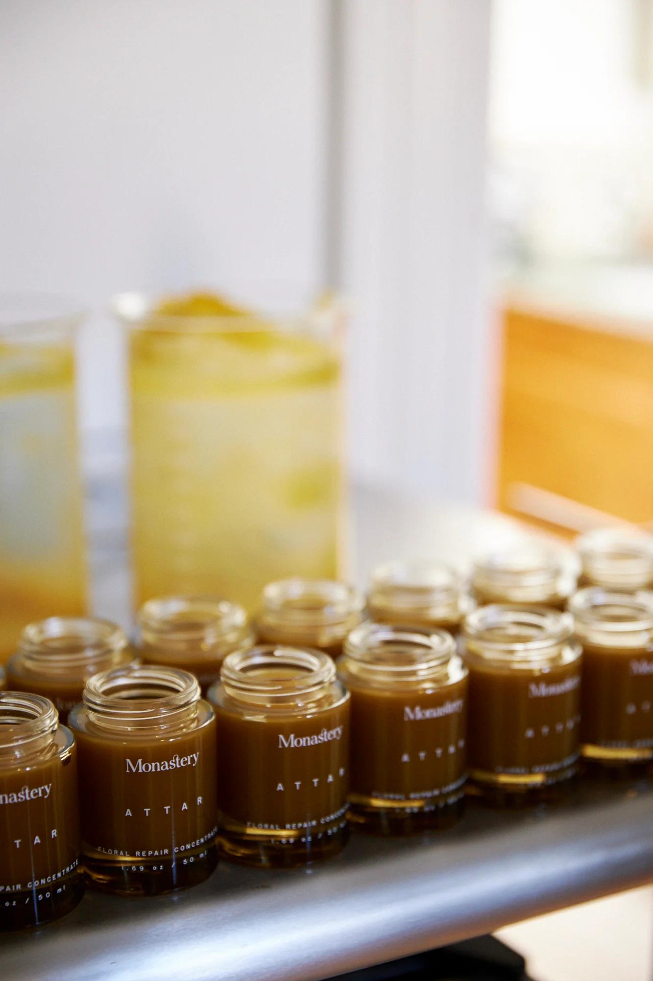 Small glass jars labeled "Monastery Attar" on reflective surface. Filled with brown liquid. Blurred yellow containers in background.