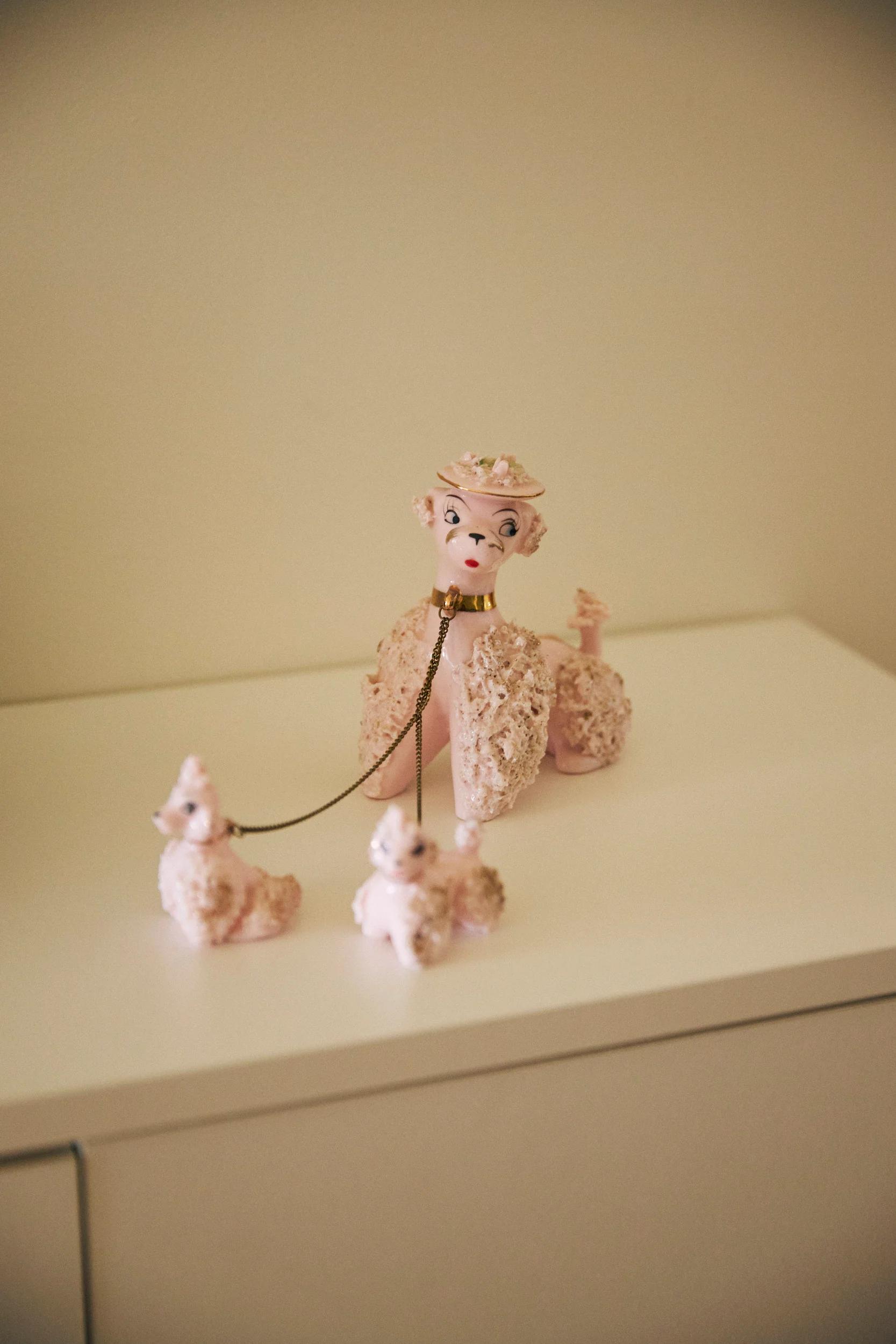 Decorative porcelain poodle figurine with pink hat on white surface. Attached to smaller poodles by chain leash.