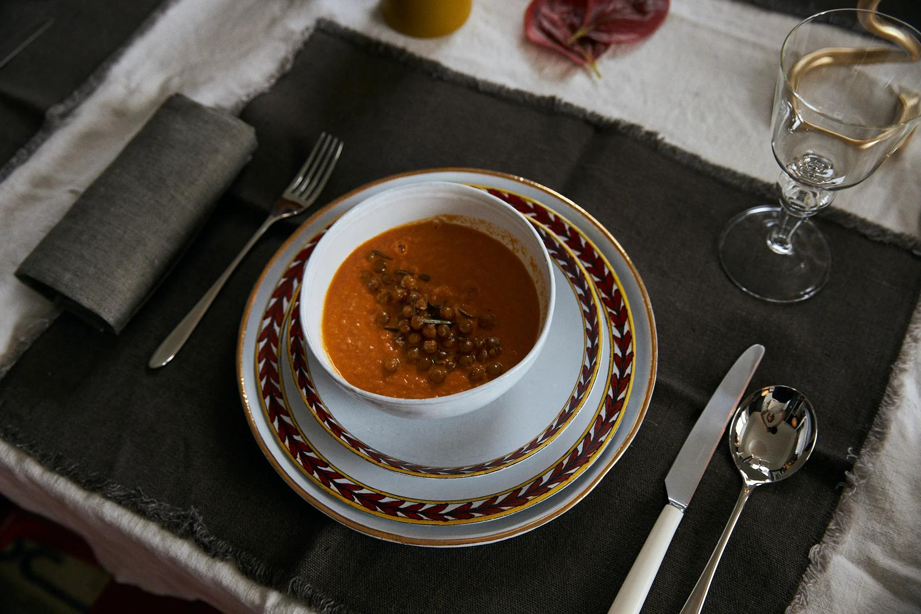 A soup bowl on decorative plates with geometric patterns. Cutlery is neatly arranged on a dark placemat, with a napkin and wine glass nearby.