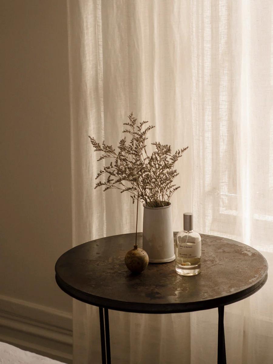 Serene Parisian vignette: Round metal table with white vase of dried flowers, Miller et Bertaux perfume bottle, and decorative object. Softly lit sheer curtain background.