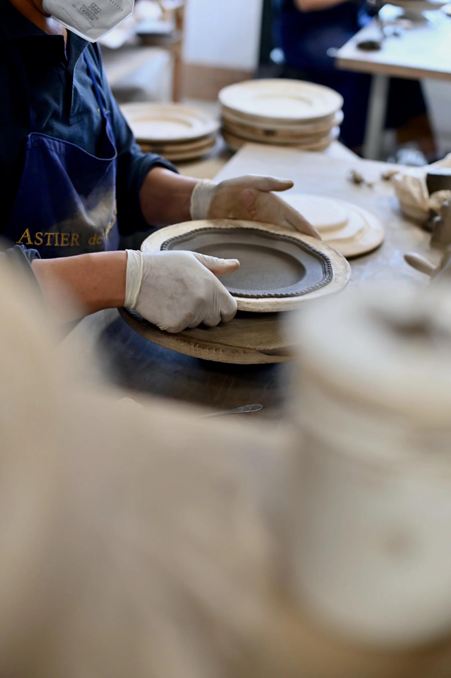 Person wearing gloves forming ceramic plate on pottery wheel. Finished and unfinished plates in background. Face partially obscured by mask.