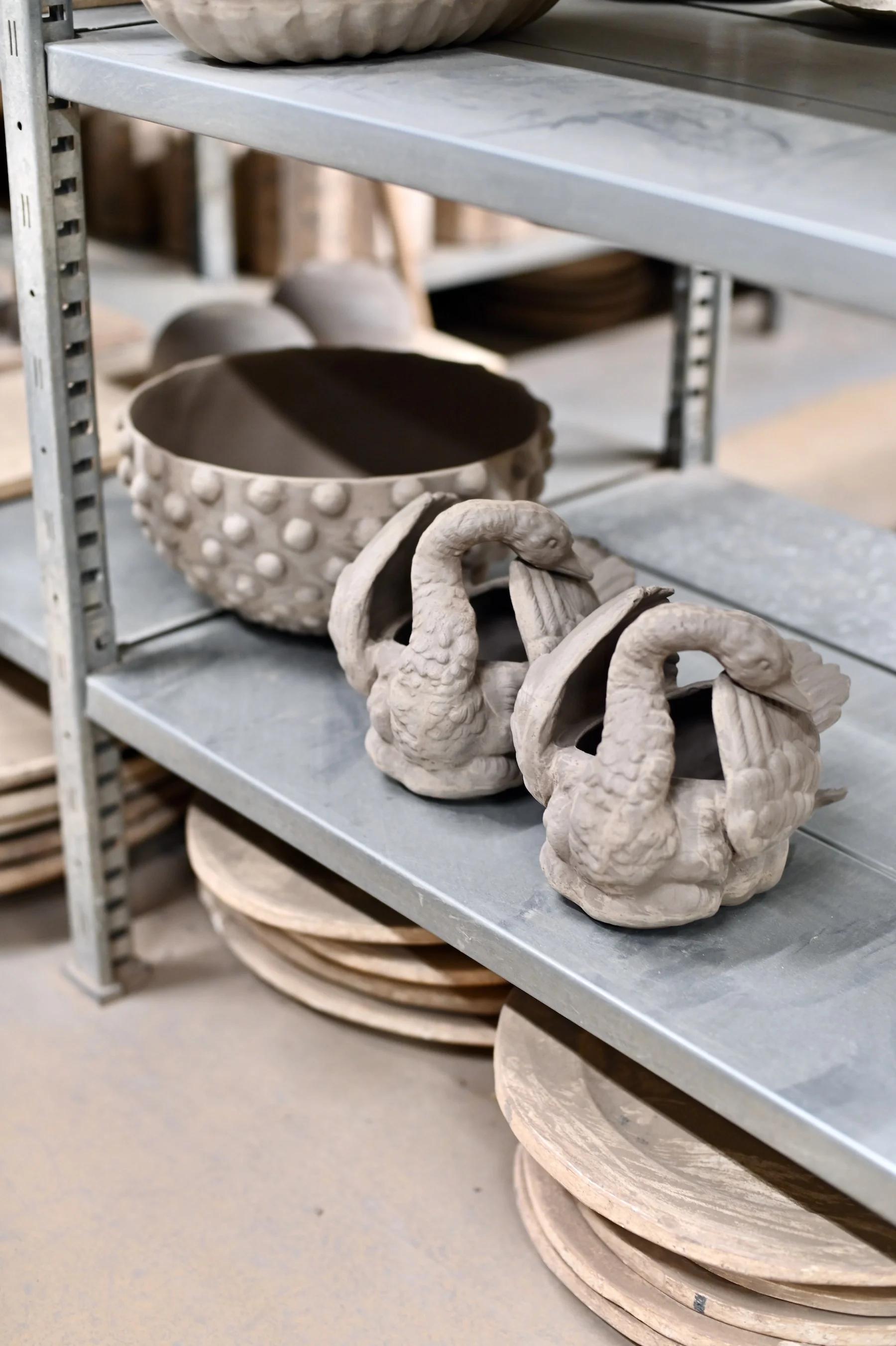 Metal shelf displays clay pottery in various stages. Two swan-shaped containers on middle shelf with large textured bowl. Bottom shelf holds stacked round plates.