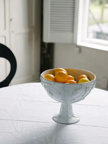 White pedestal bowl of lemons on round table near window. Soft light illuminates scene with glimpses of black chair and white cabinets.