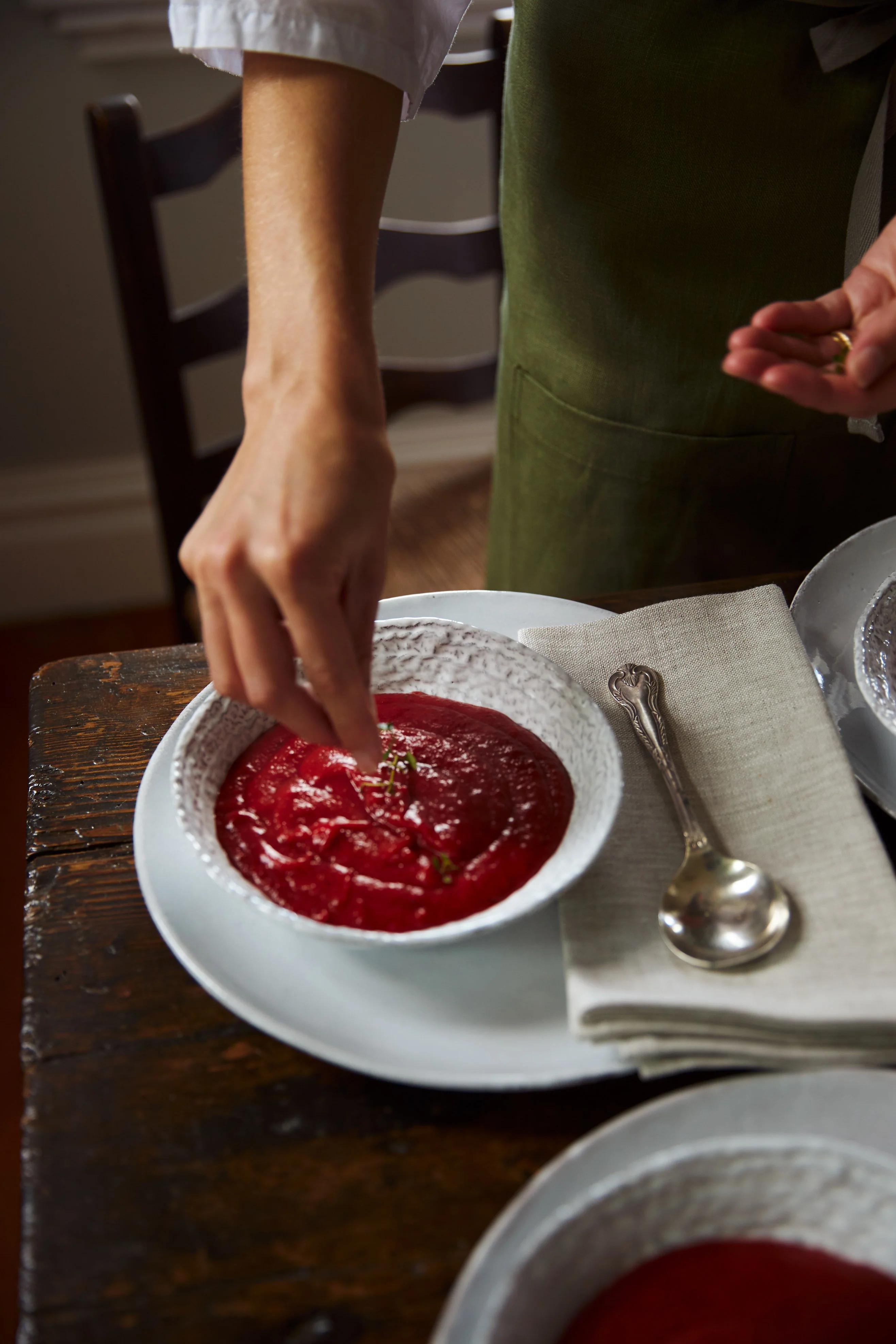 A person sprinkles herbs onto a bowl of red beetroot soup on a white plate, with a spoon and napkin beside it.