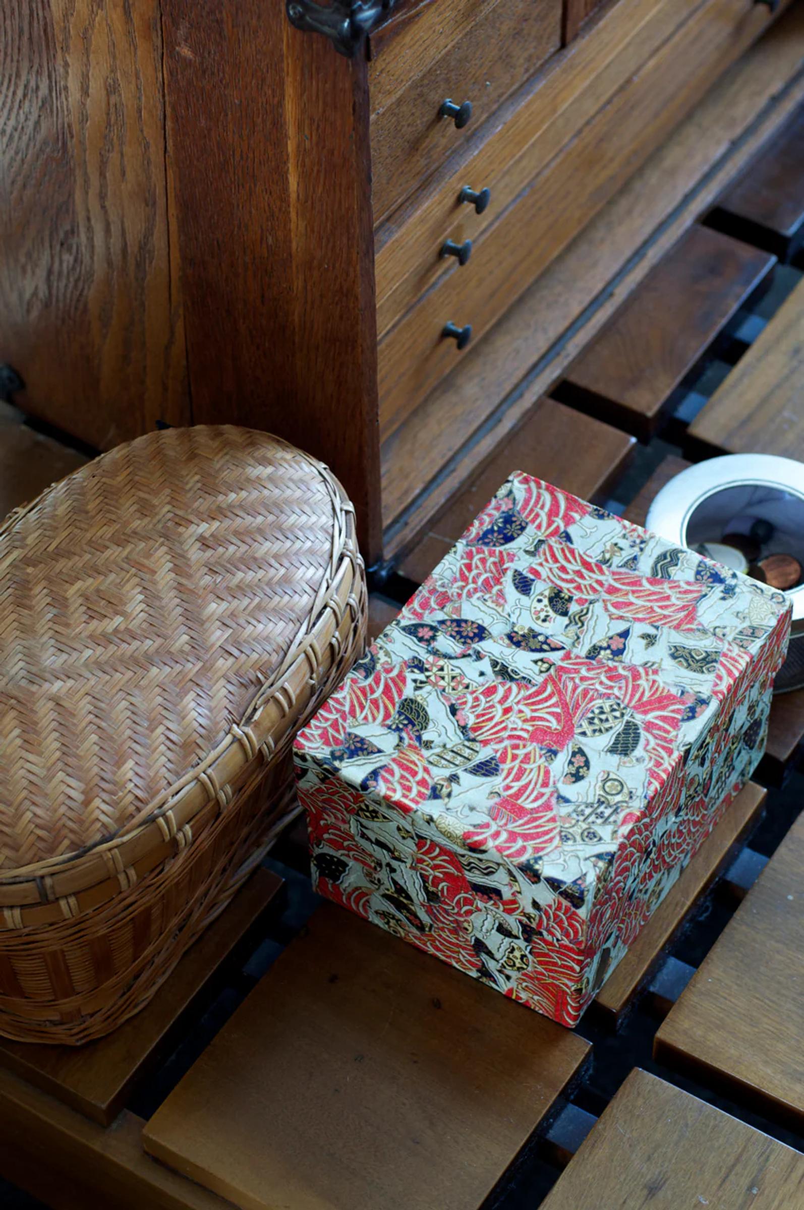 Woven oval basket and decorative box with cat pattern on wooden surface. Tape roll in frame. Wooden cabinet with drawers in background.