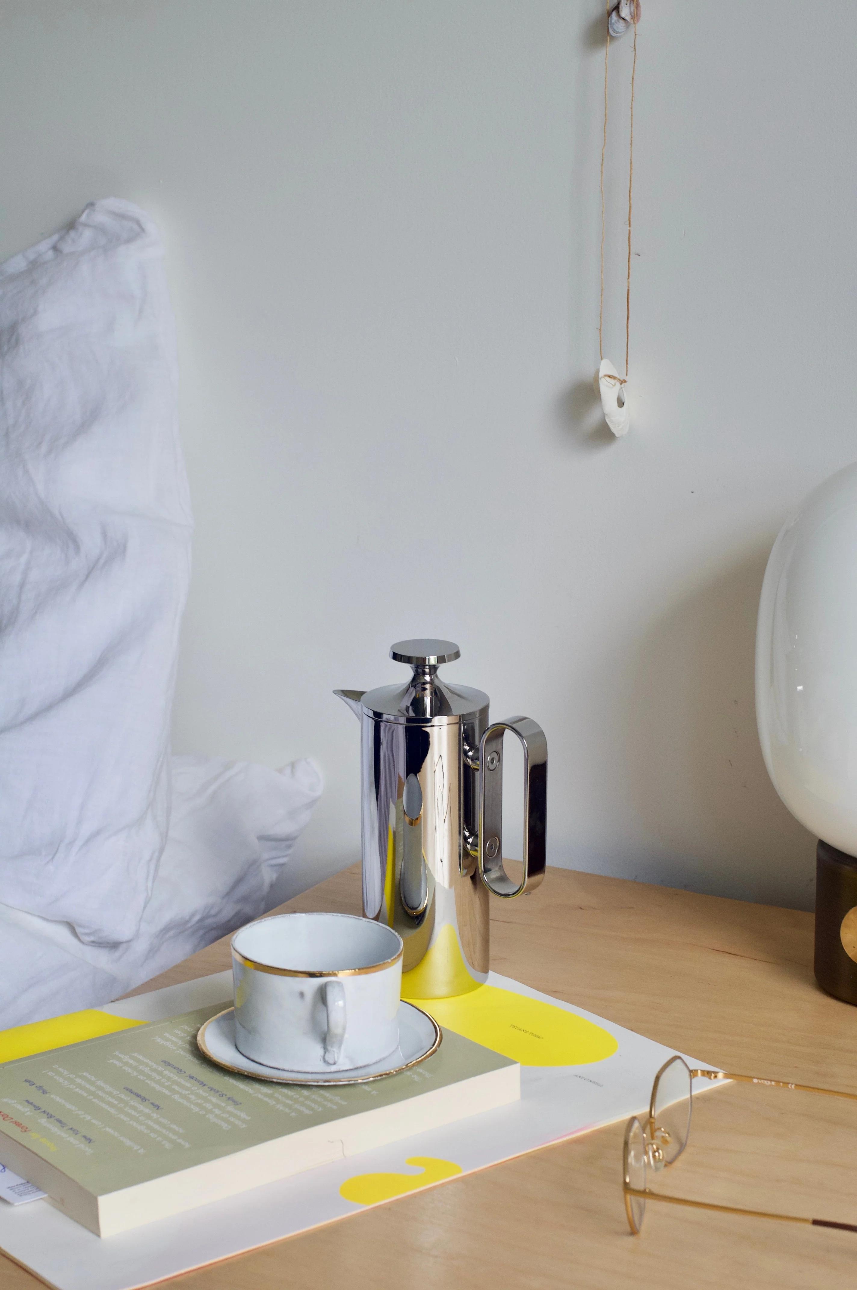 Cosy bedroom with white-sheeted bed, wooden nightstand holding David Mellor cafetière, teacup on book, glasses, and modern lamp. Wall decoration visible.