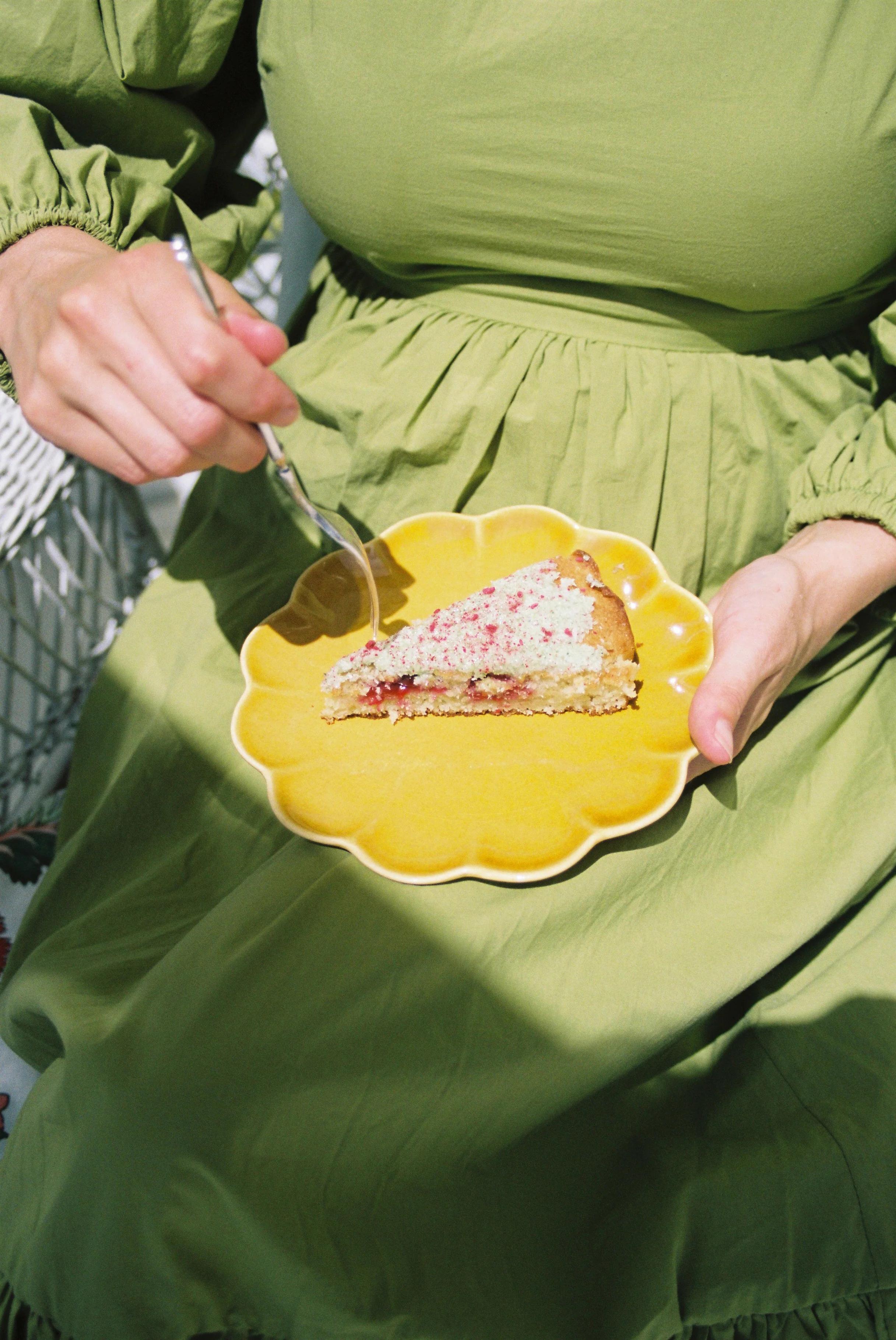 Overhead view: Person in green dress eating cake slice from yellow flower-shaped plate with fork.