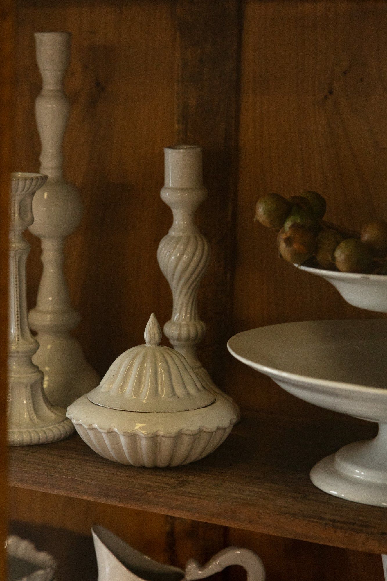 Wooden shelf with white ceramic items: candle holders, lidded dish, tall candlestick, and pedestal plate with green pods.