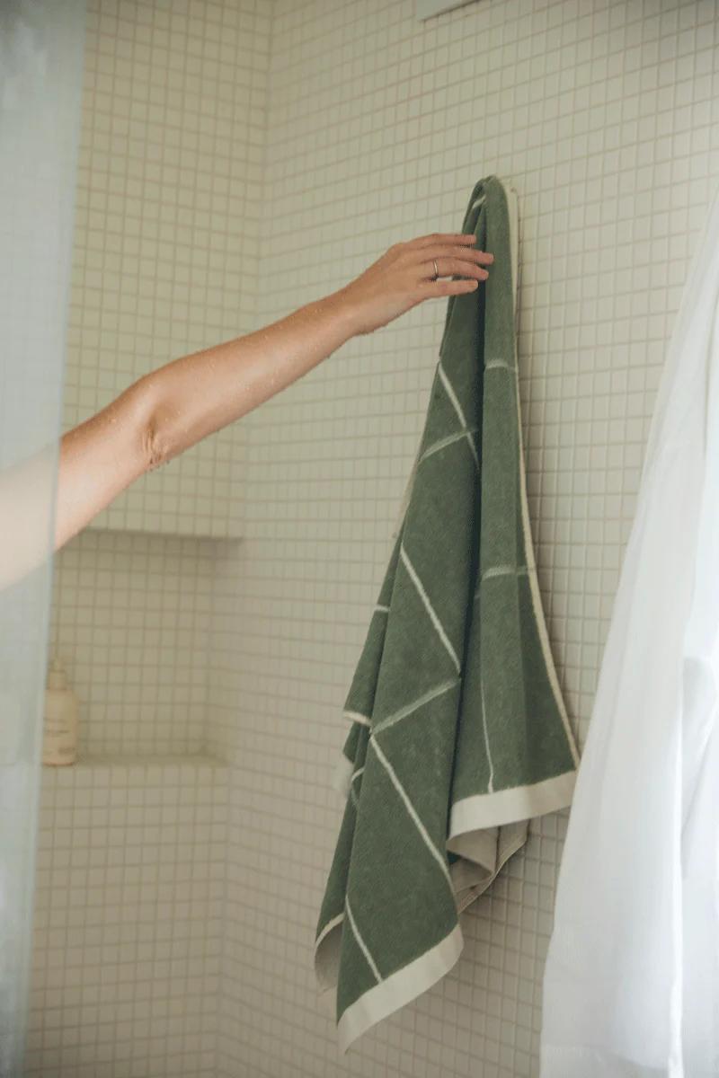 A hand reaches out from a shower to hang a green geometric-patterned towel on a hook. A white bathrobe hangs nearby against white tiled walls, reflecting Baina’s craftsmanship.