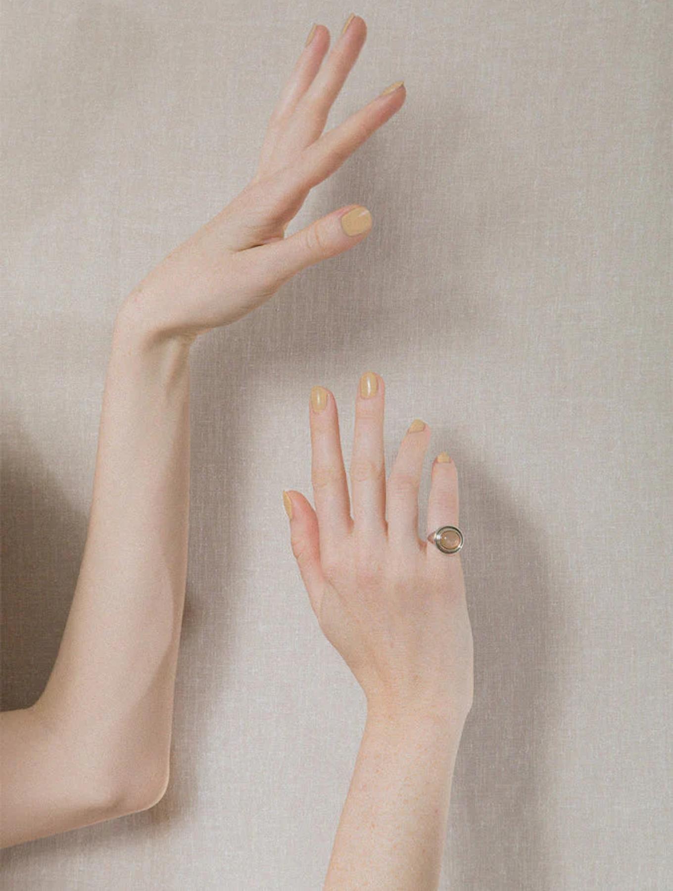 Two fair-skinned hands against beige background. Left hand raised, right hand lower with ring. Both display nude nail polish.