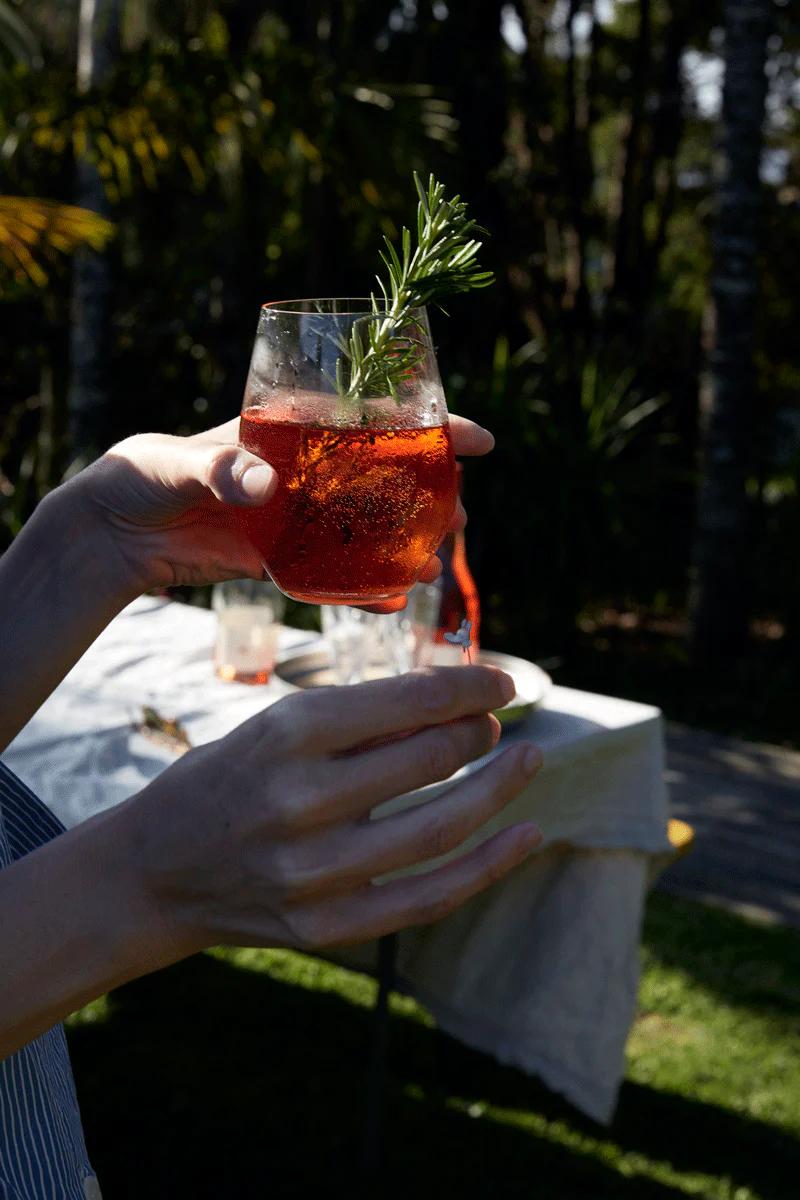 Person holding red drink with rosemary sprig at garden party. White-clothed table and greenery in background.