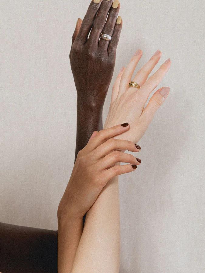 Three intertwined hands reaching upward, each in a different tone. The light and dark tones wear rings, and all three are wearing nail polish.