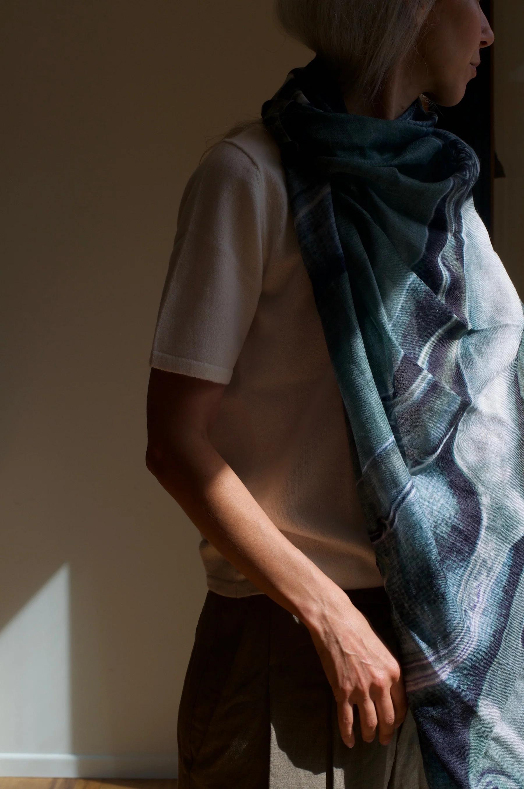 Long-haired person in light shirt and patterned Good & Co scarf. Standing in partial shadow with soft natural light highlighting arm and torso.