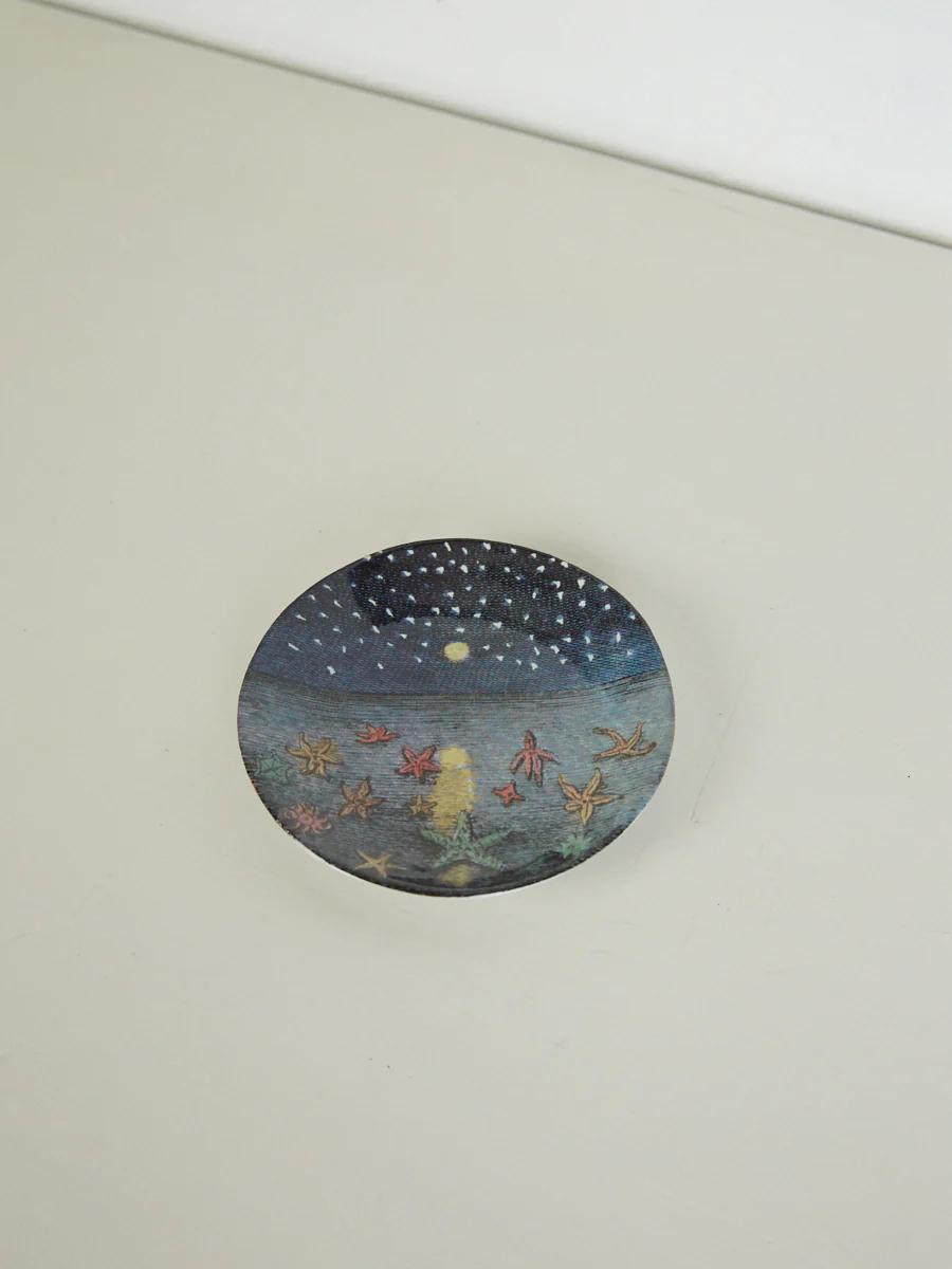 Round dish with night sky design. Stars, crescent moon. Yellow and red shapes, water reflection. Light surface.