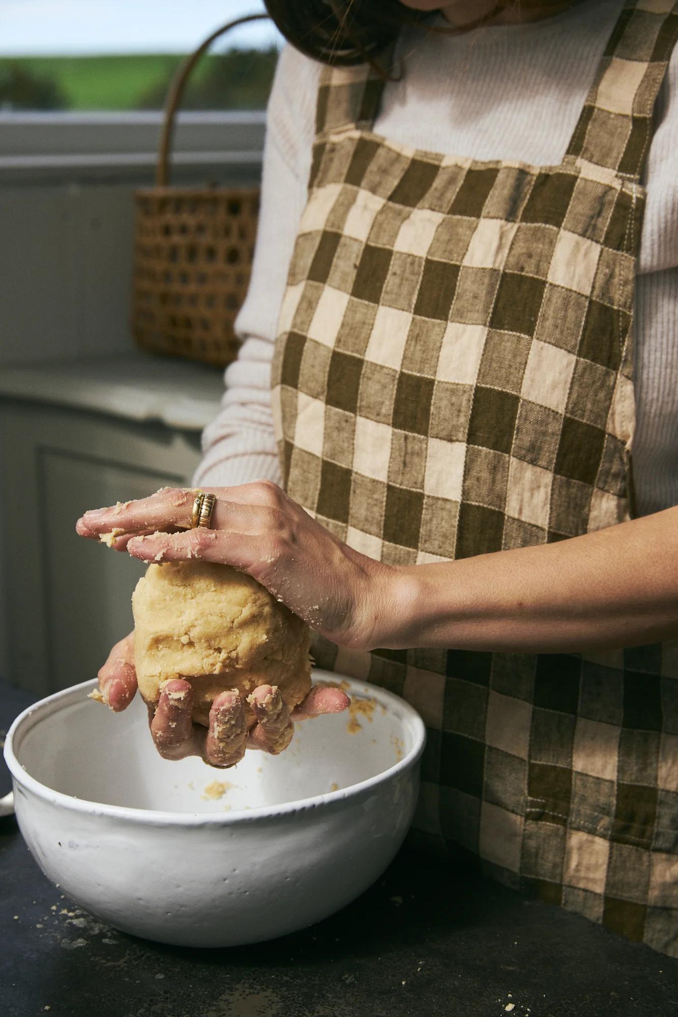 Person in checkered apron kneading dough over white bowl. Wicker basket with fruits near window.