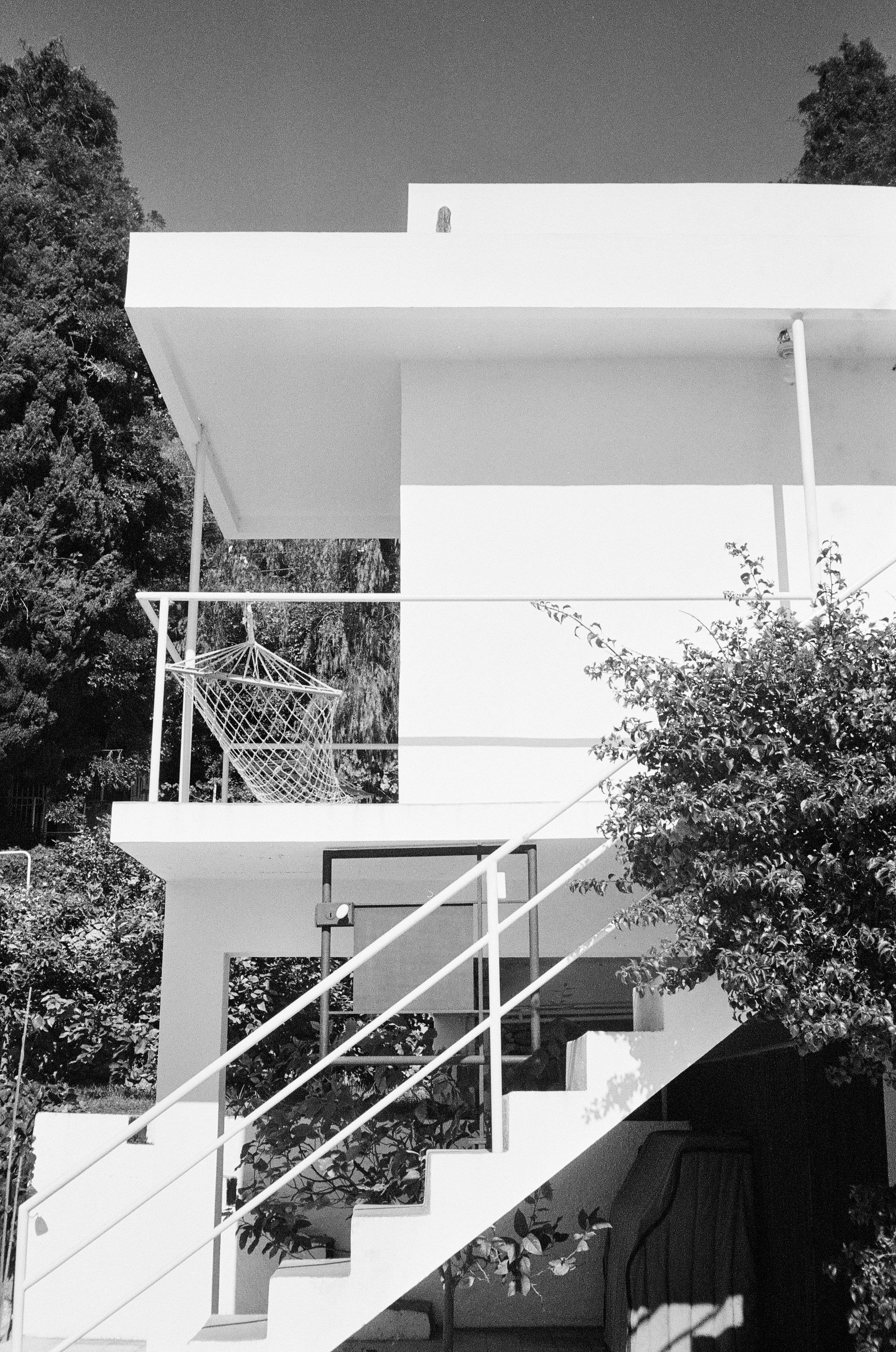 Black and white photo of modernist two-story house. Exterior staircase leads to balcony with hammock chair. Trees surround house.