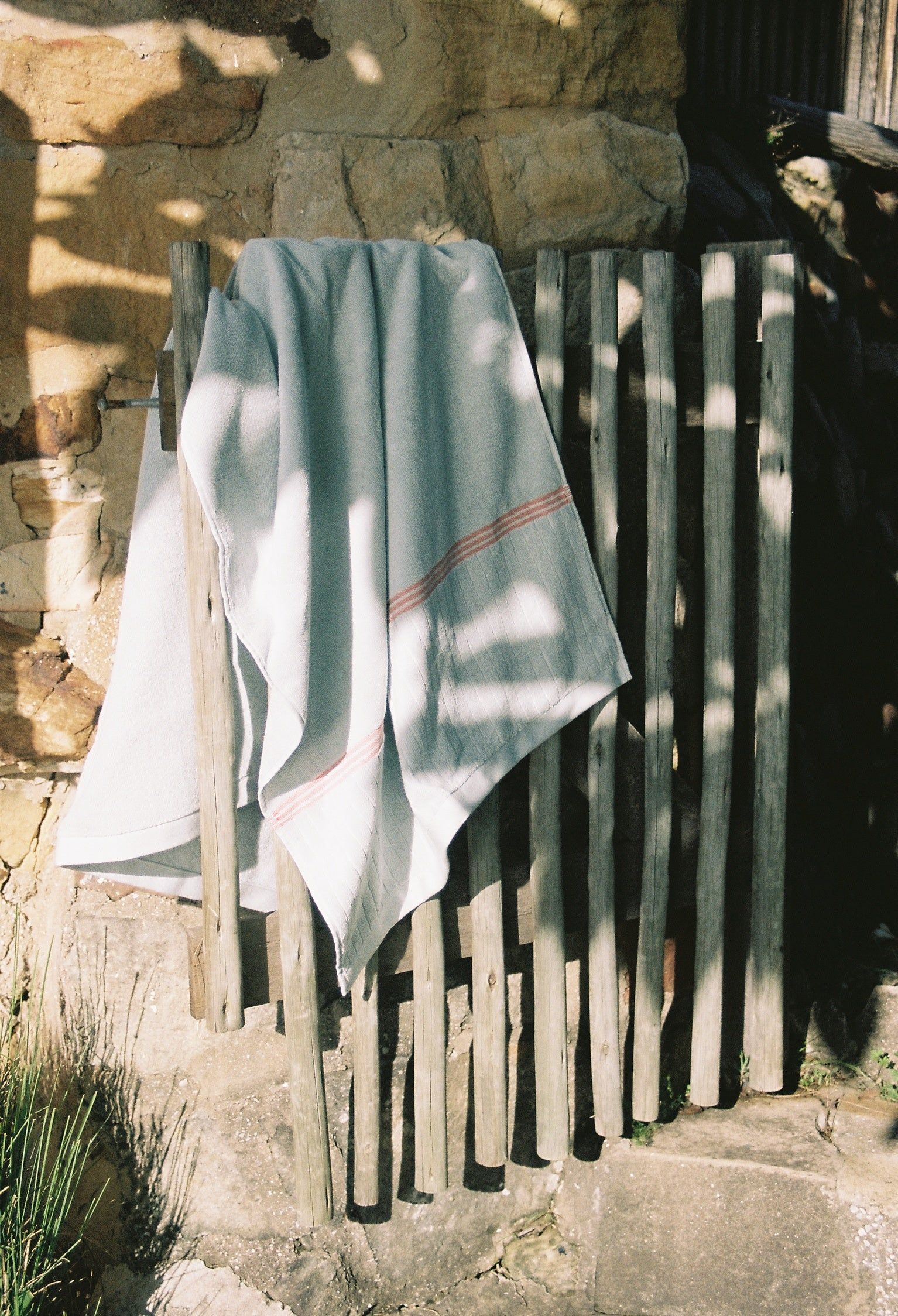 A light blue towel with red stripes hangs over a rustic wooden fence, partially in shadow, with a stone wall and greenery in the background.