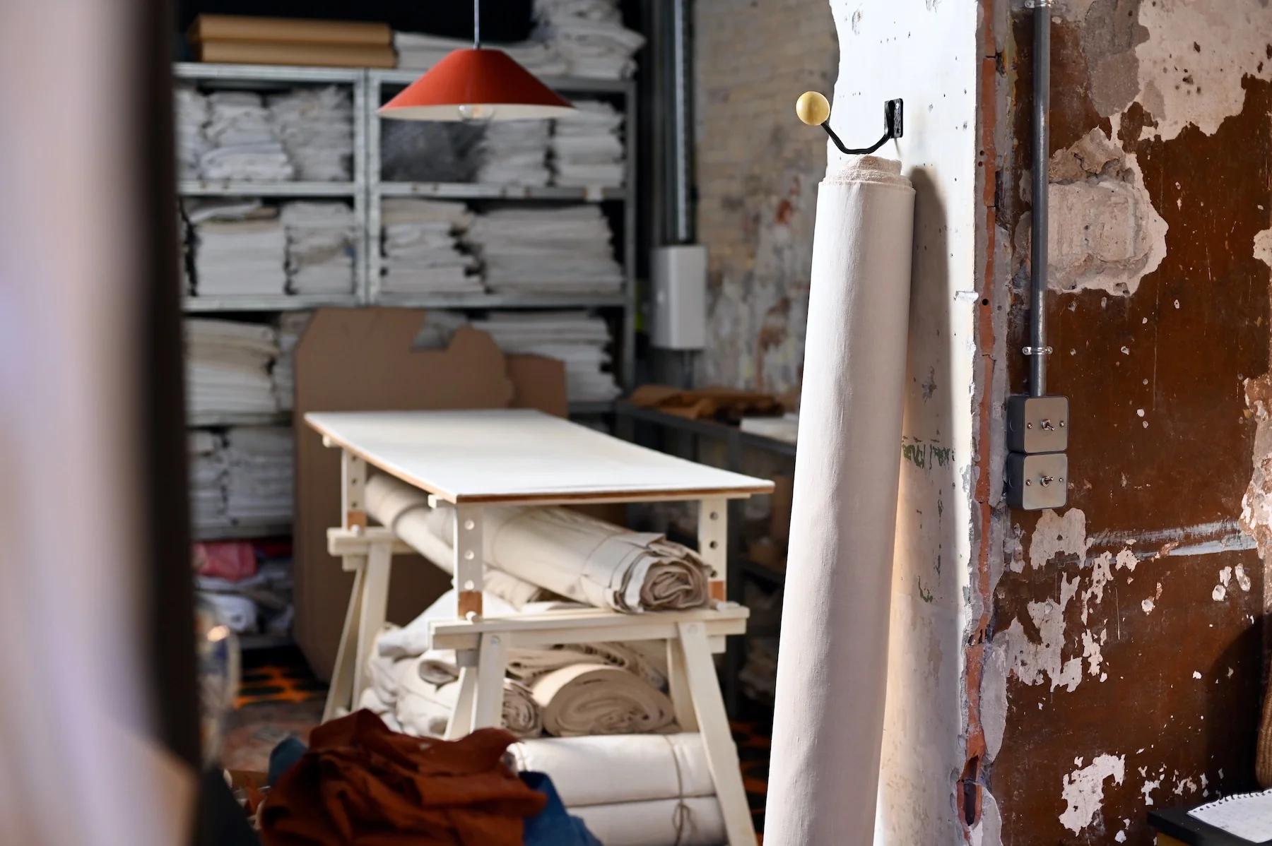 Room with fabric-filled shelves and central table with fabric rolls. Red lampshade hangs above. Worn walls with peeling paint.