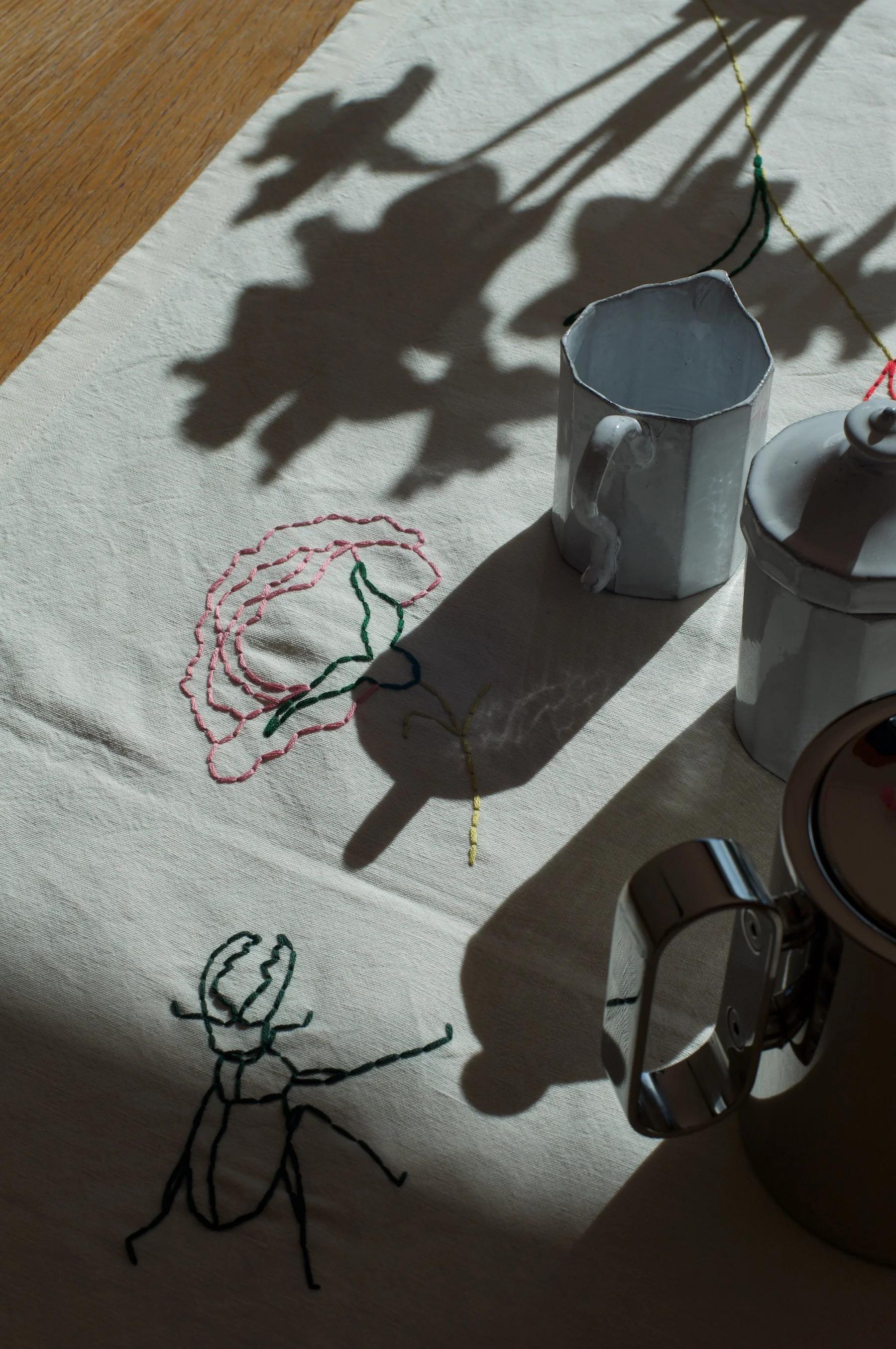 Table with embroidered cloth featuring flower and beetle. Metal cups and small pitcher. Natural light casting shadows.