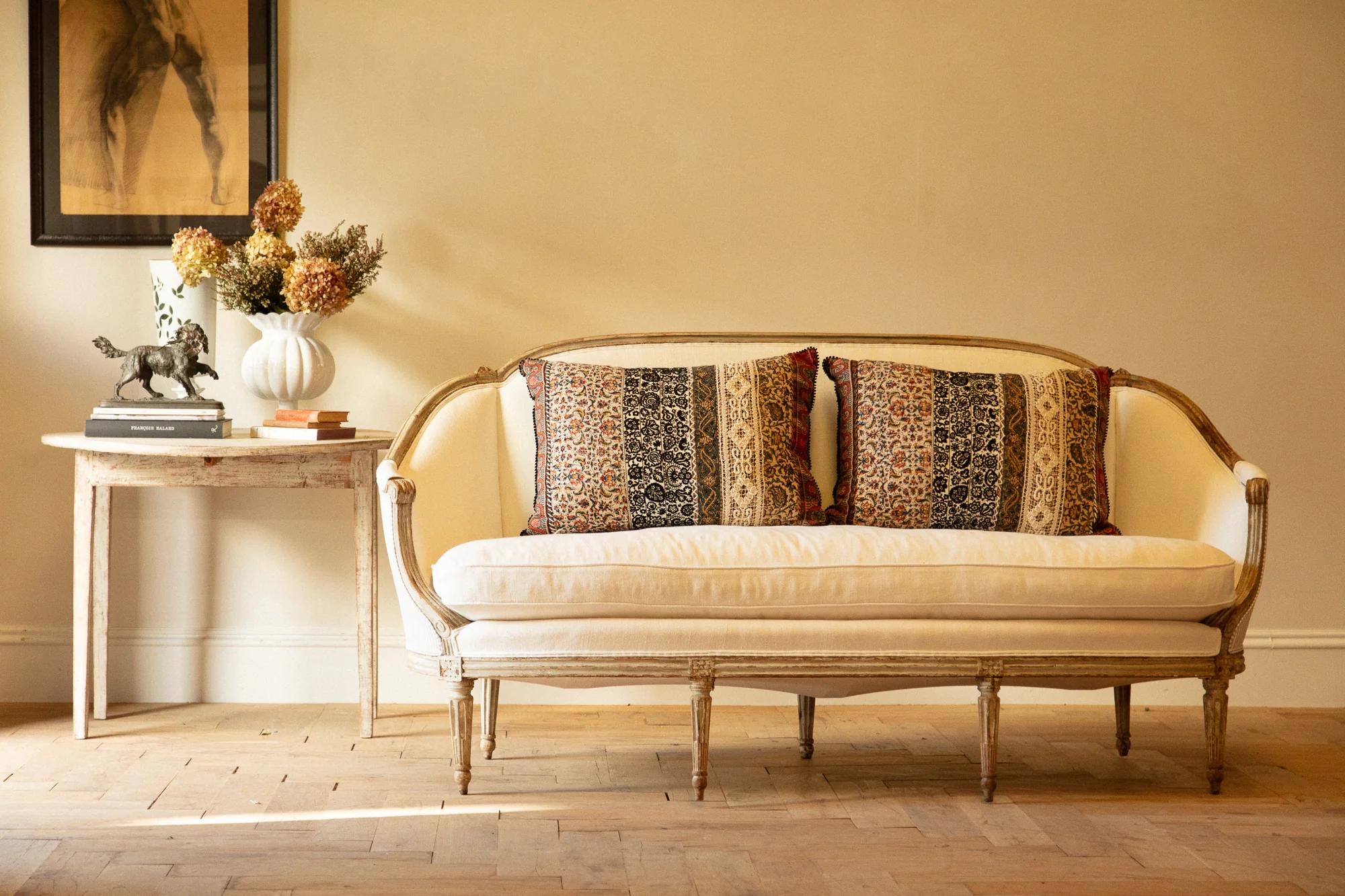 Beige loveseat with patterned cushions. Side table holds vase, horse sculpture, books. Horse painting above. Wooden floor pattern.