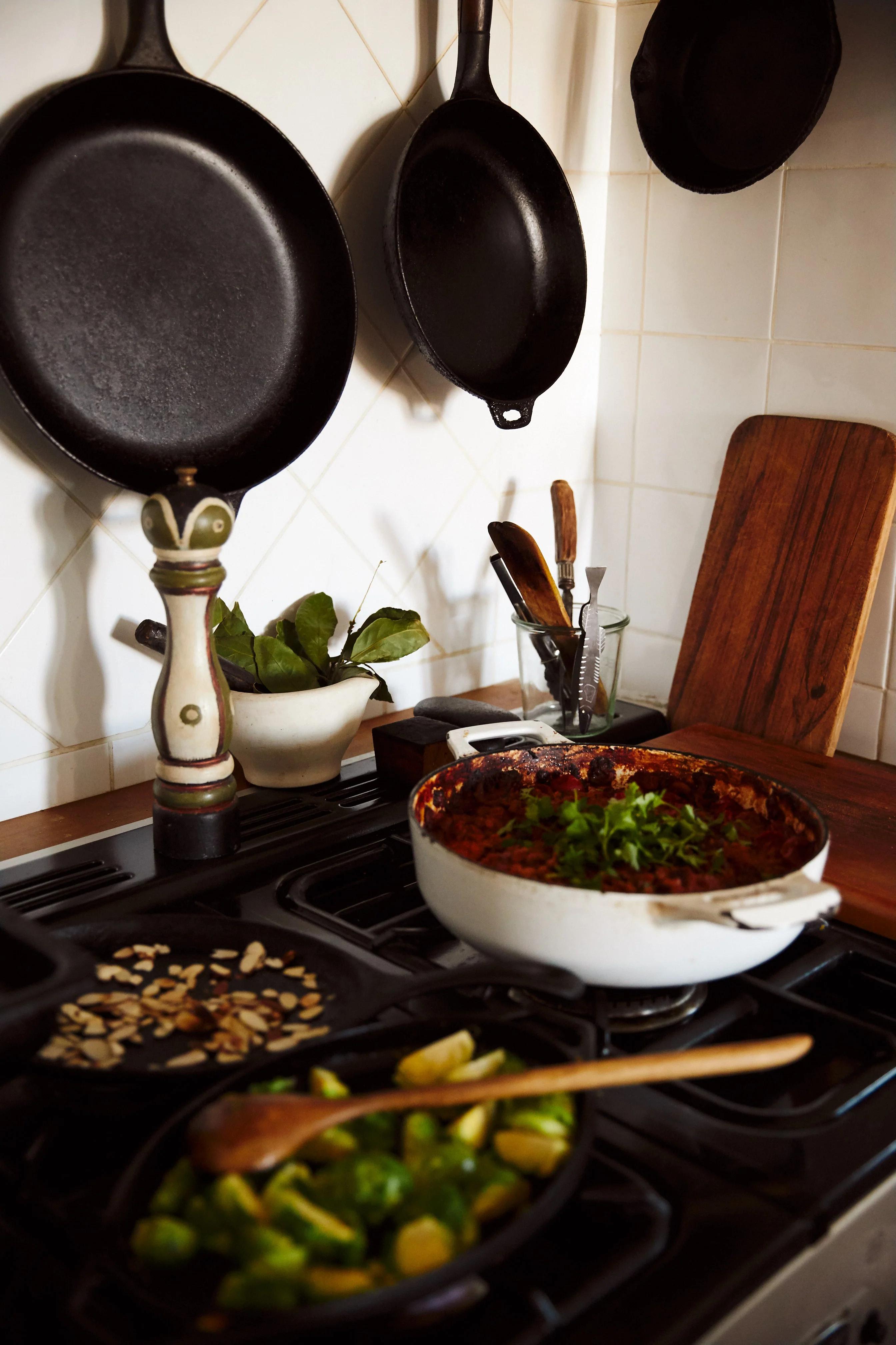 Stovetop with herb-garnished dish and sautéed vegetables. Cast iron skillets hanging above.