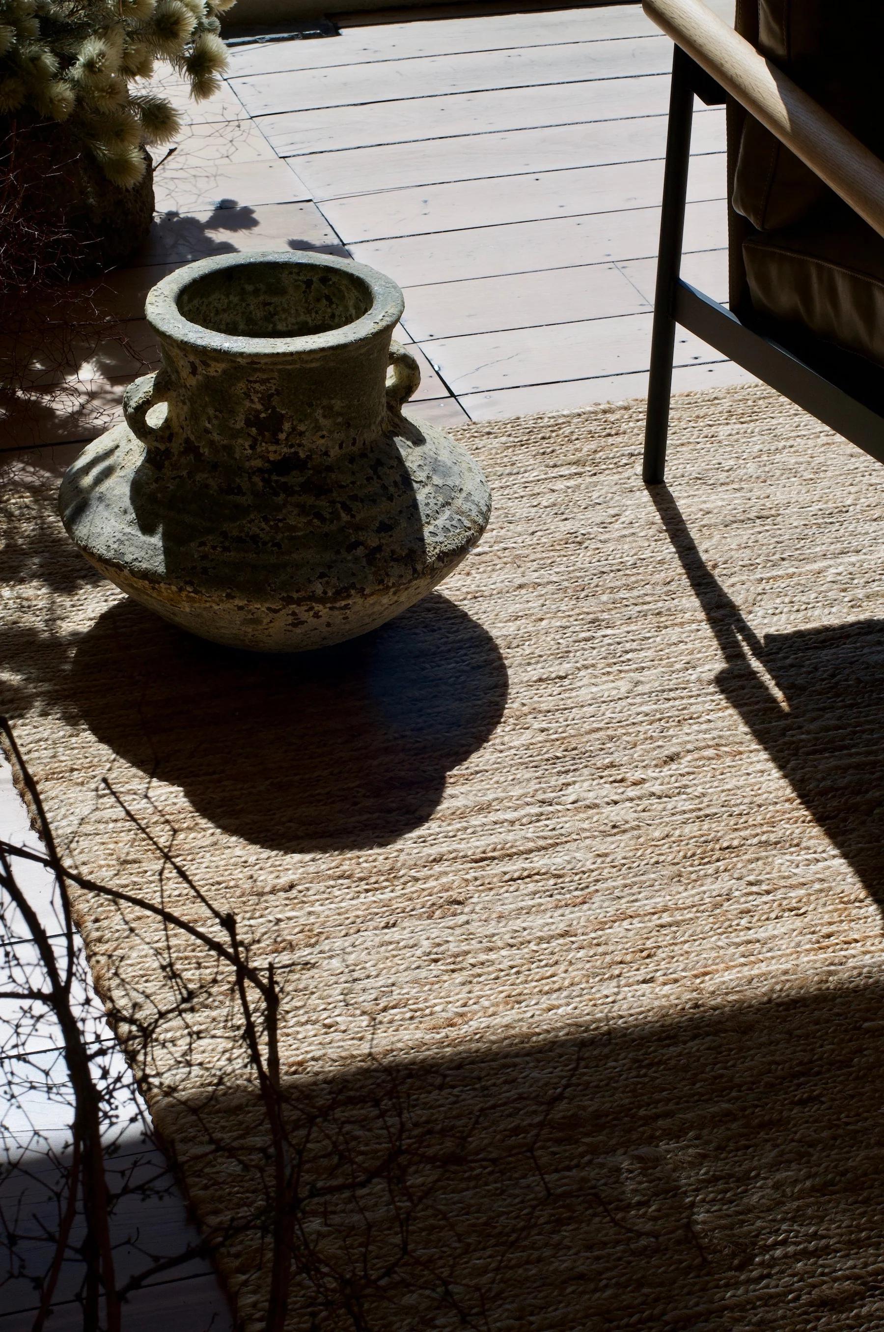 Ceramic pot casting shadow on sunlit rug. Wooden chair and plants visible, creating rustic atmosphere.