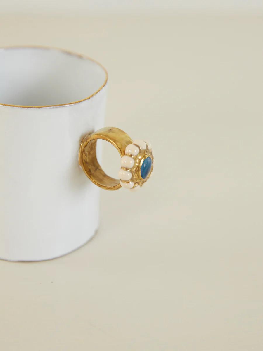 White ceramic cup with gold rim and ornate ring handle. Blue stone surrounded by pearls. Neutral beige surface.