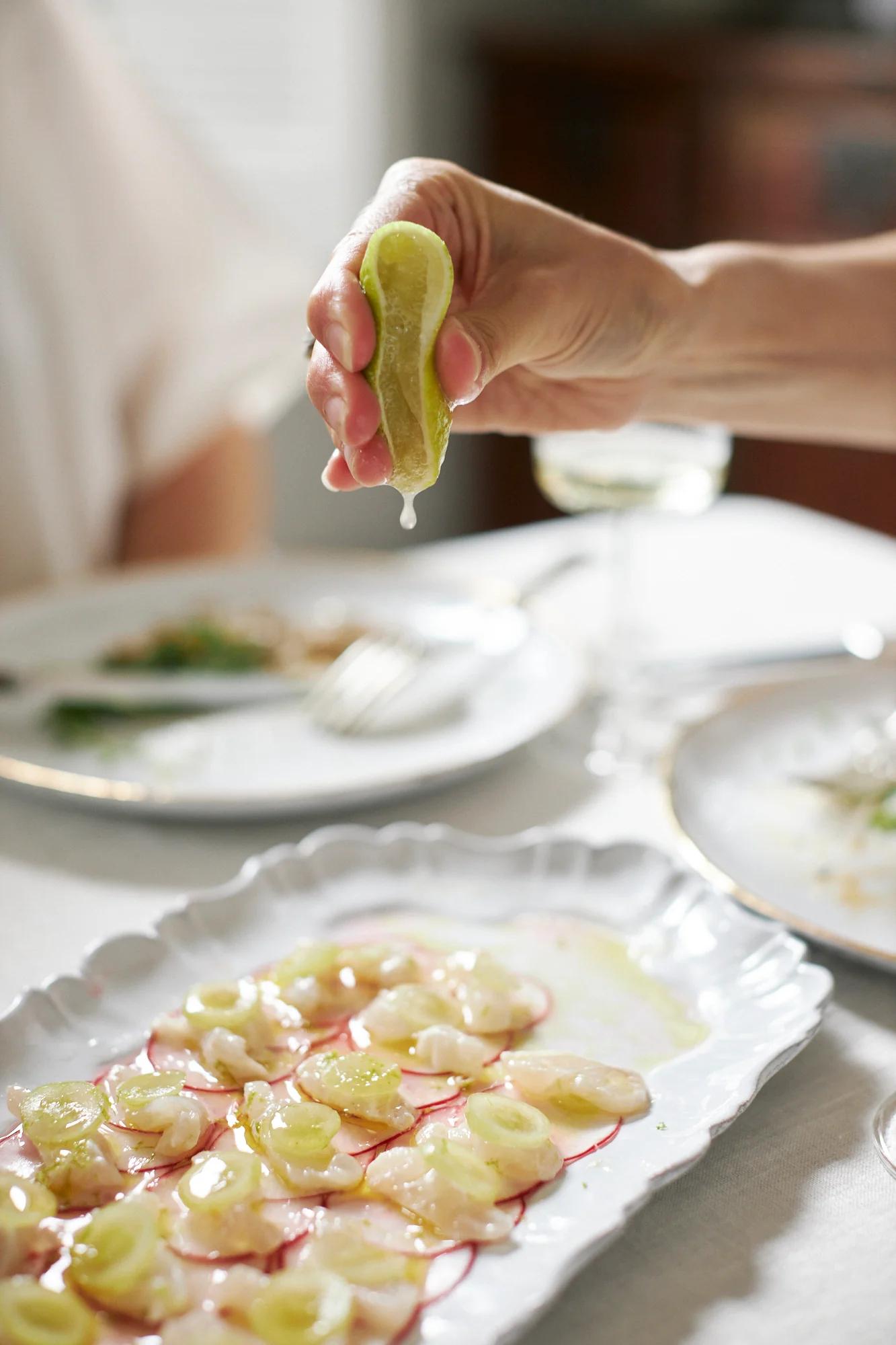 A hand squeezes lime over seafood and radishes on a white dish. Other plates and a wine glass are visible on the white tablecloth.