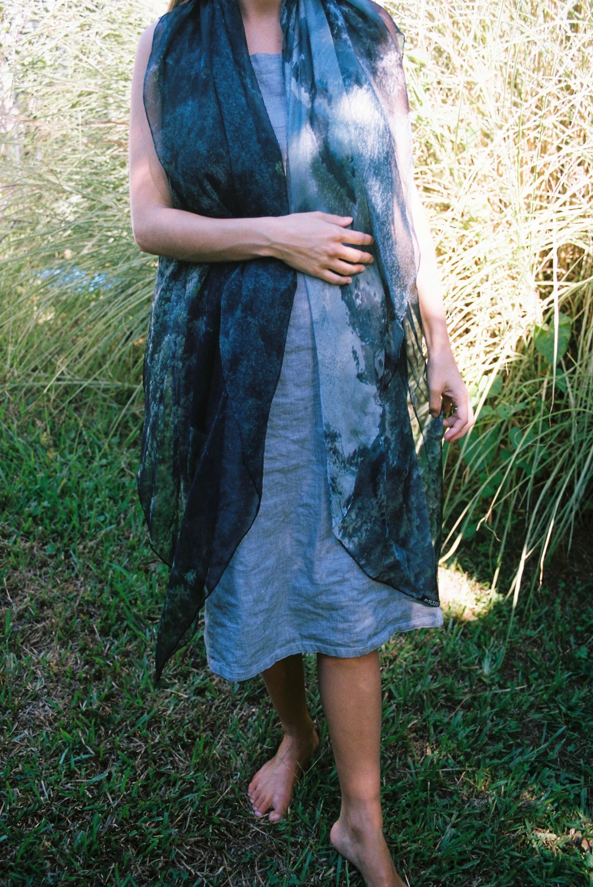 Barefoot person in light grey dress with large, dark patterned scarf. Standing on grass with foliage background.