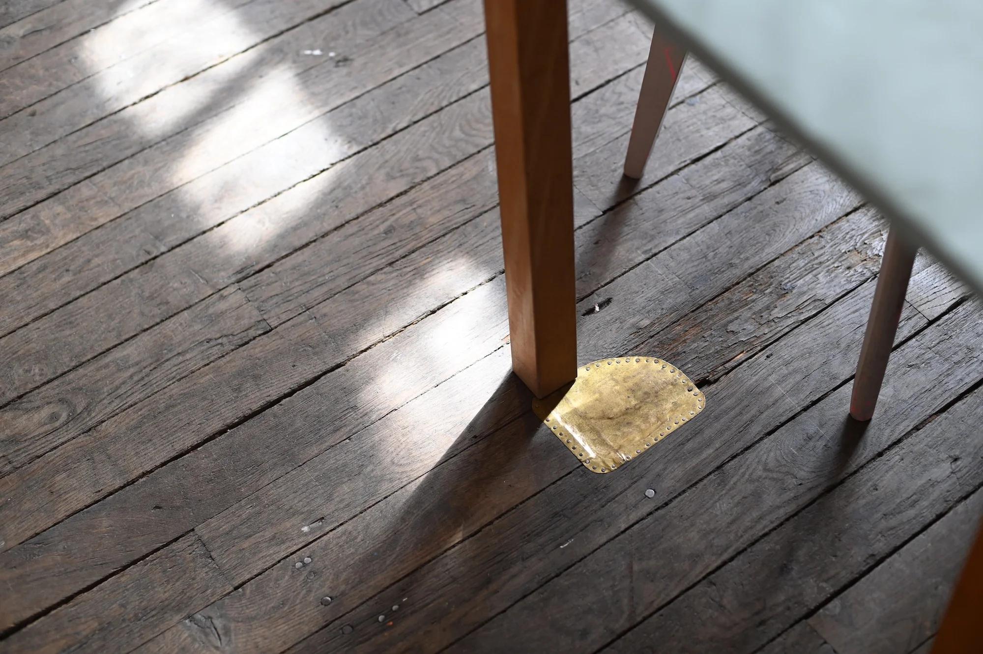 Close-up of wooden floor with gold metal patch. Table leg nearby. Light creates elegant shadow pattern.