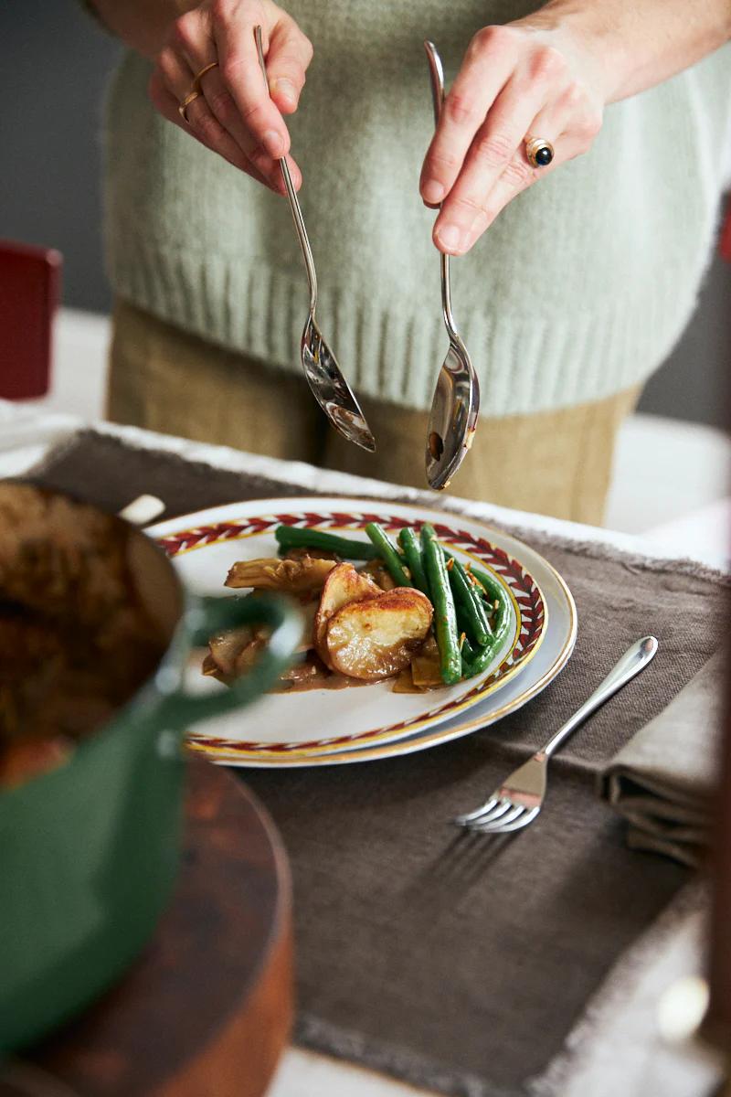 Person serving roasted vegetables onto decorative plate. Green pot of beef casserole partially visible. Grey tablecloth.