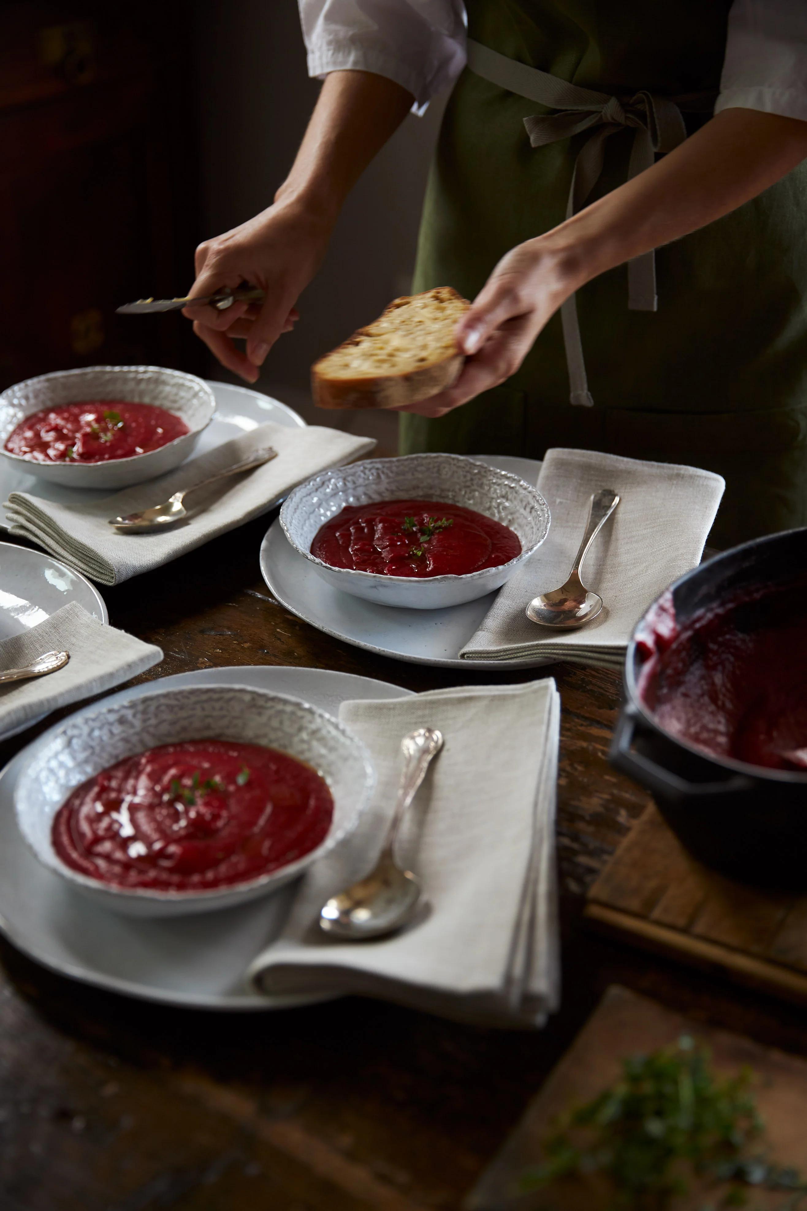 A person in a green apron serves bread alongside three bowls of beetroot soup, each on a napkin with a spoon. The three visible bowls are garnished with herbs.