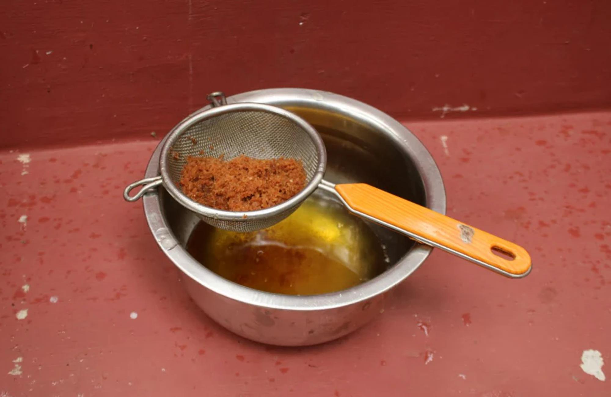 Metal bowl with yellow liquid on maroon surface. Metal strainer with orange handle and brown residue over bowl.